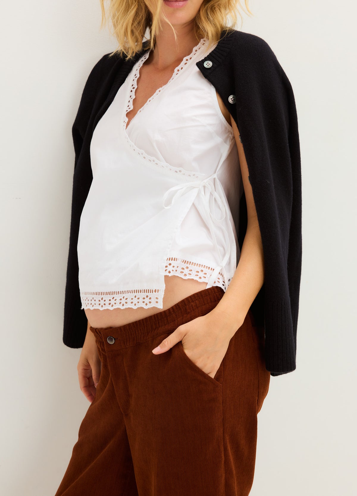 White lace-trimmed maternity nursing top with side tie, paired with brown pants and black cardigan draped over shoulders.