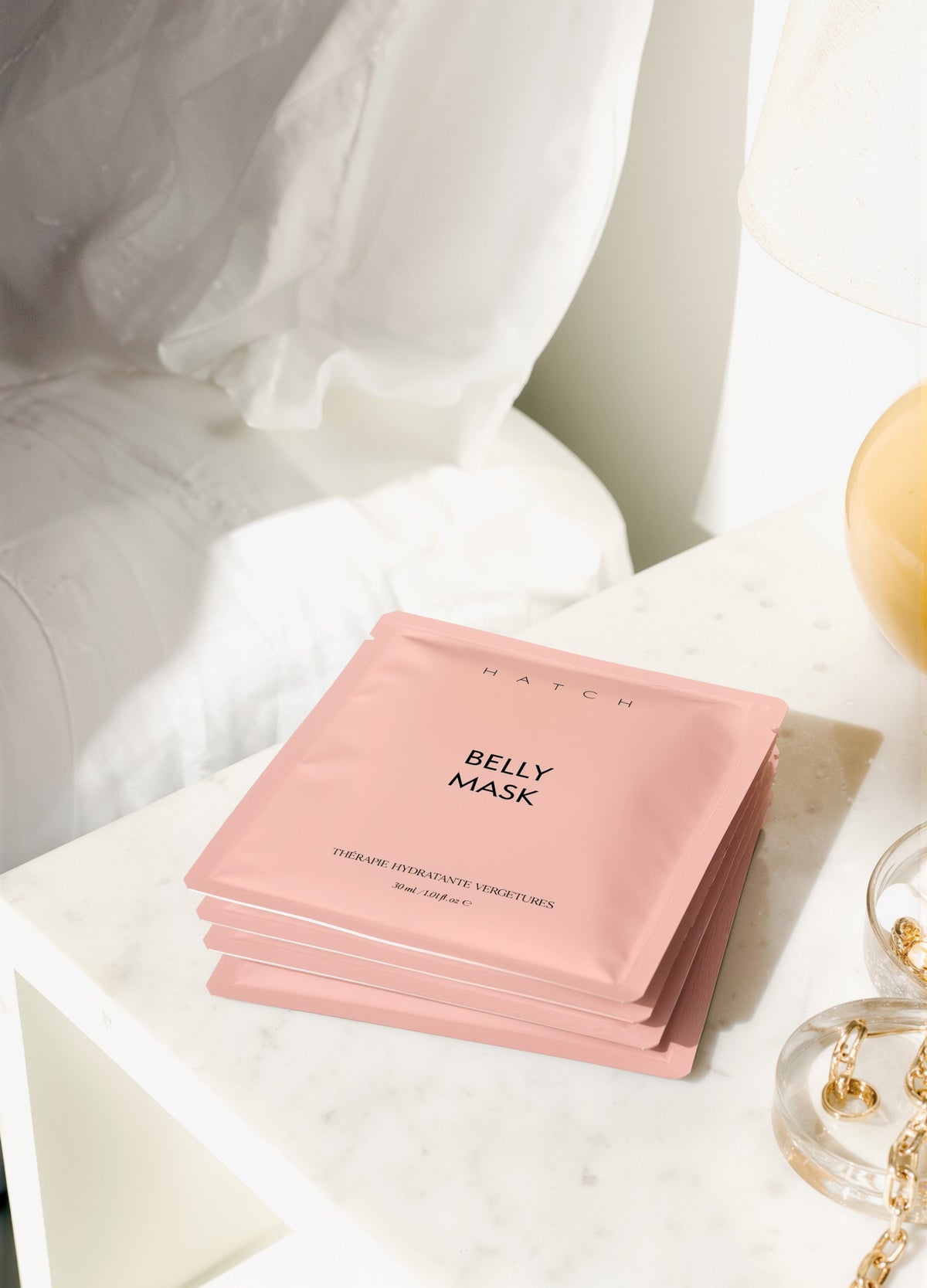 Stack of matte pink Belly Mask Pack sachets with simple black text on a white bedside table.