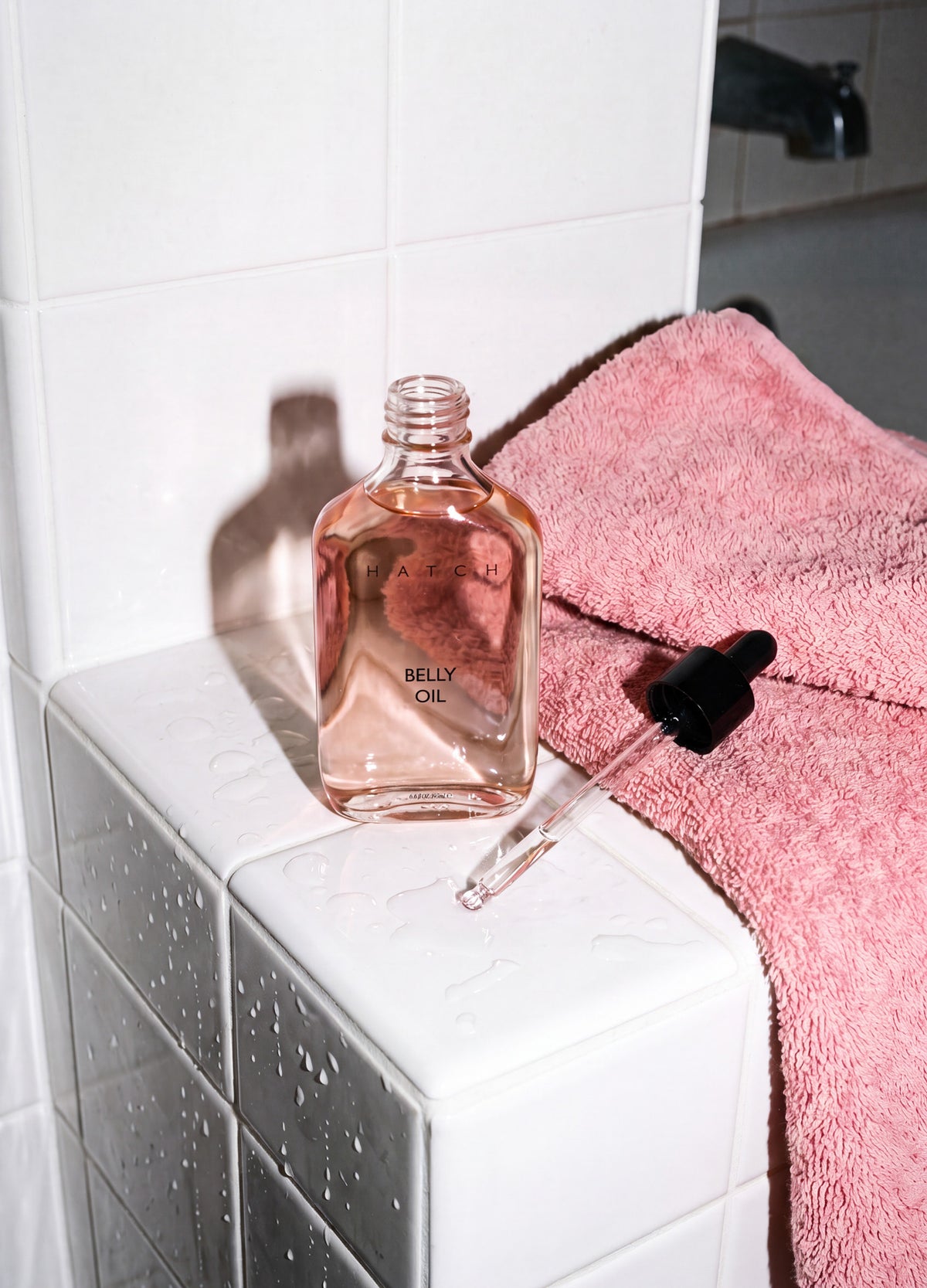 Clear rectangular glass bottle with light pink belly oil and black dropper cap on white tiled surface next to soft pink.