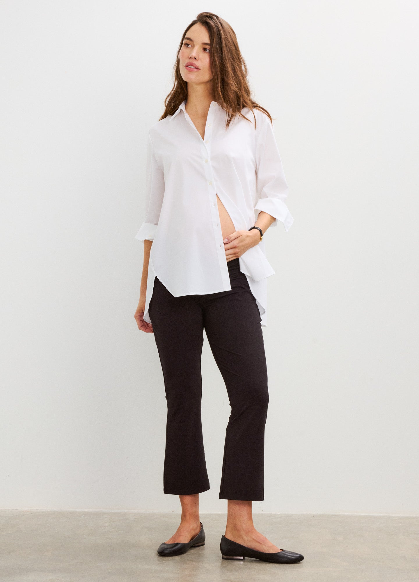 Black crop flare maternity leggings with a stretchy fit worn with a white button-up shirt and black flats.