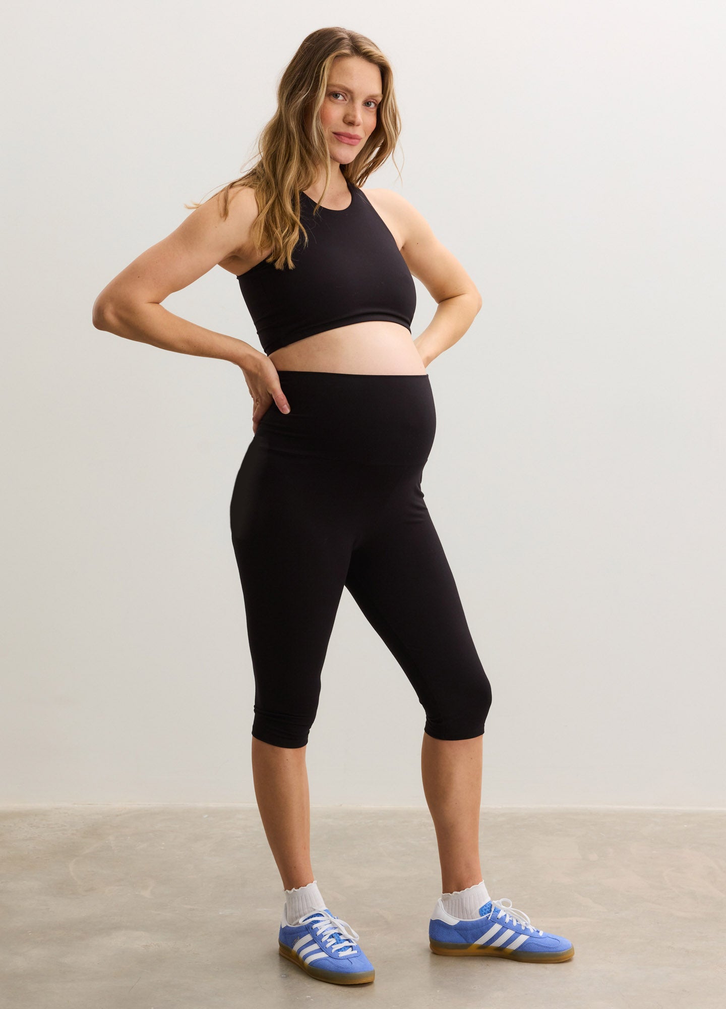 Black stretchy maternity capri leggings fitted over pregnant belly paired with black crop top and blue sneakers.
