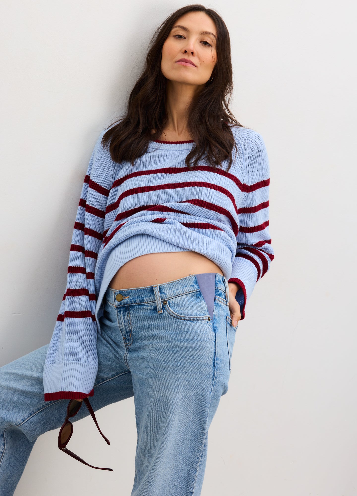 Pregnant woman wearing a blue and red striped sweater with jeans against a plain background