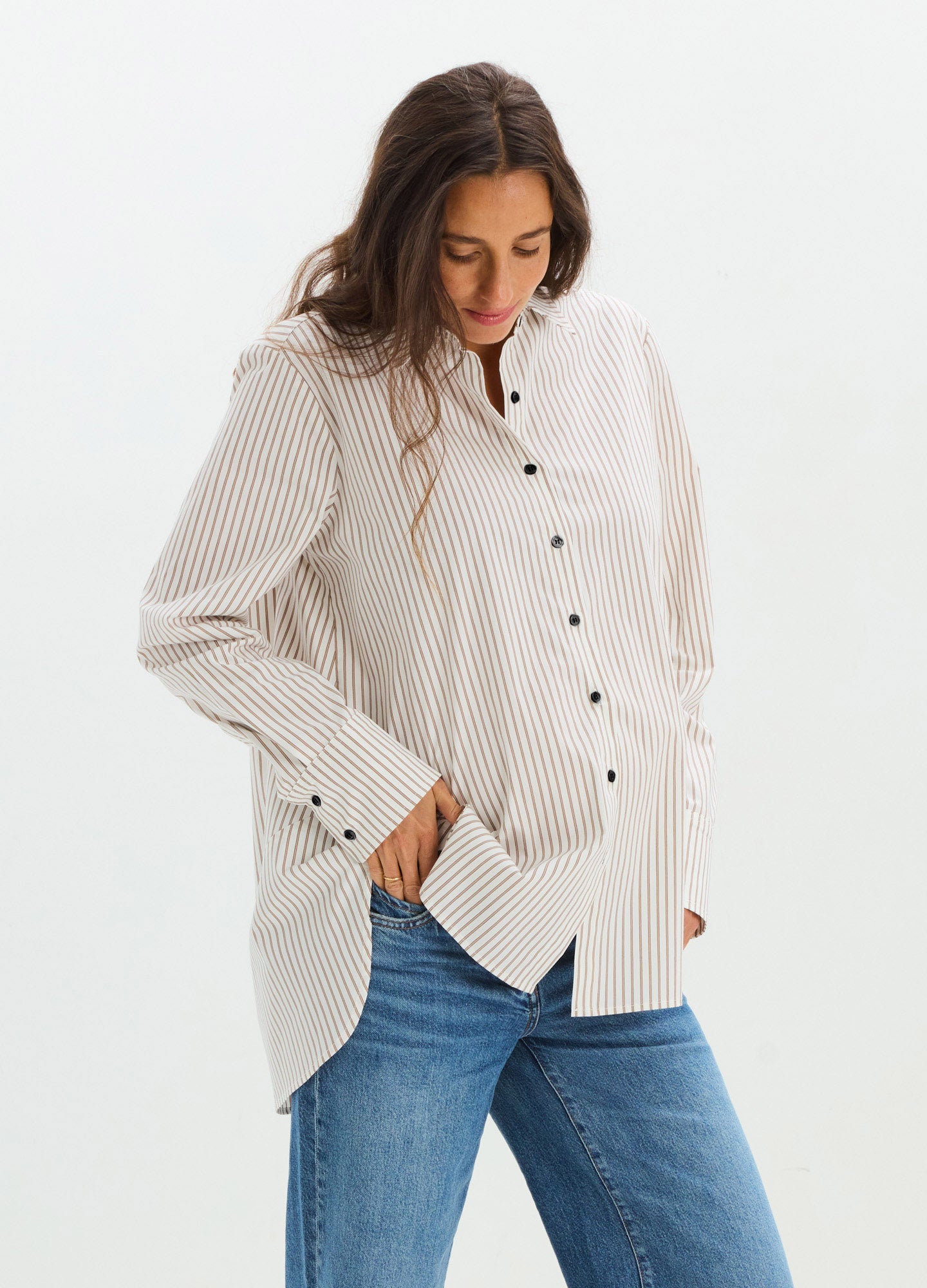 Cream and white striped loose-fit maternity button-down shirt with long sleeves and black buttons, worn over blue jeans.