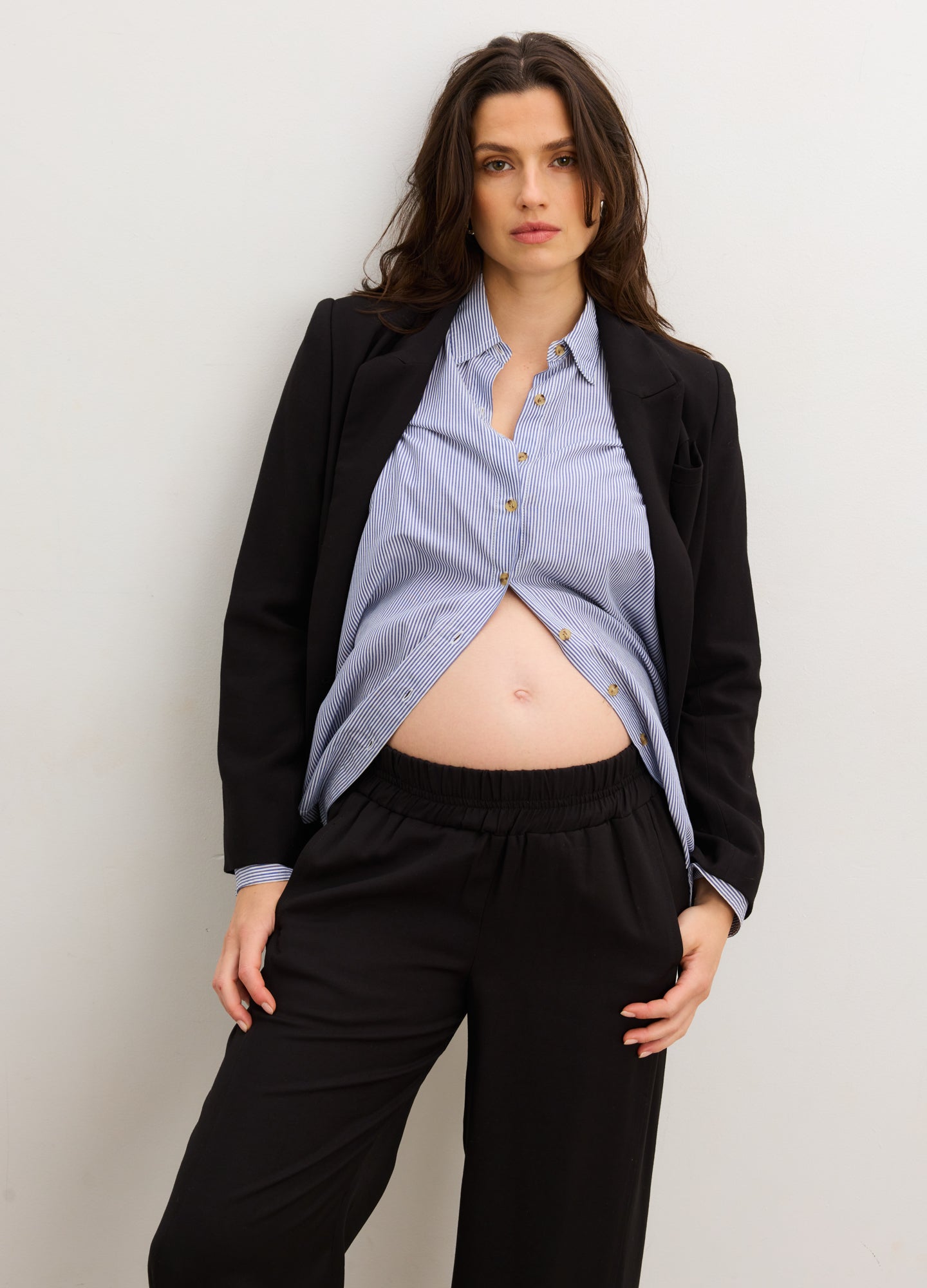 Light blue button-down maternity shirt with a curved hem, styled open over black drawstring pants and a black blazer.