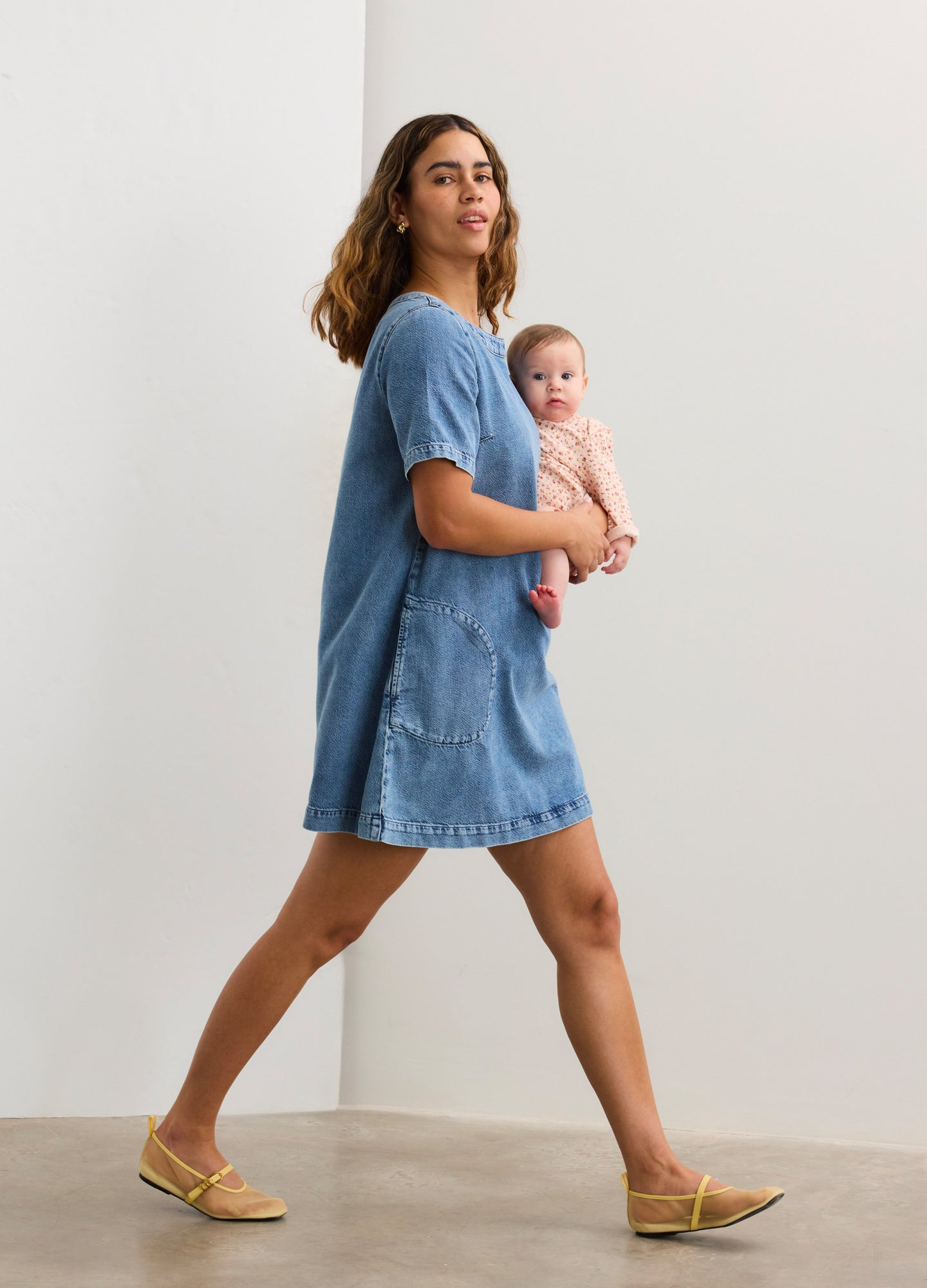 Light blue short-sleeve denim maternity dress with front pockets and relaxed fit, worn by a pregnant model holding a baby.