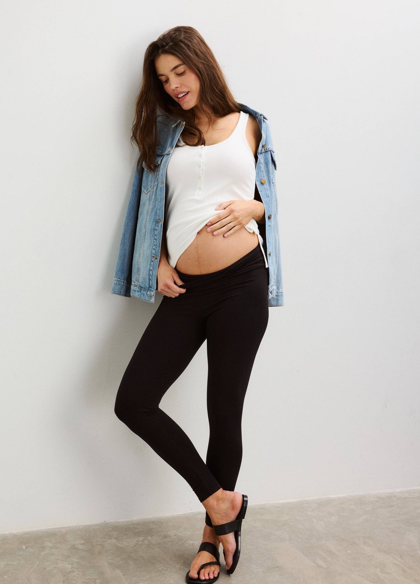 Pregnant woman wearing black maternity leggings, white tank top, and draped denim jacket with black sandals.