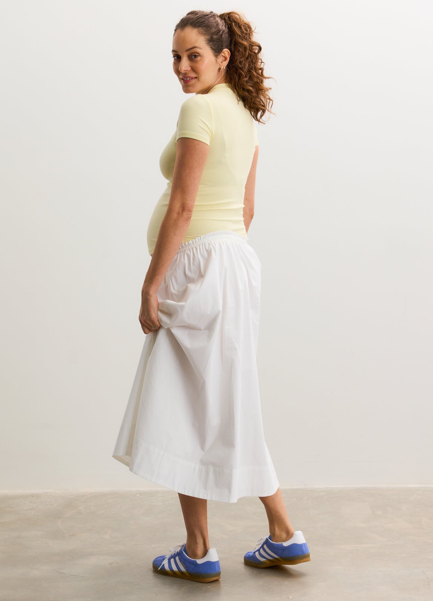 Light yellow short-sleeve ribbed maternity tee paired with white flowy midi skirt and blue sneakers.