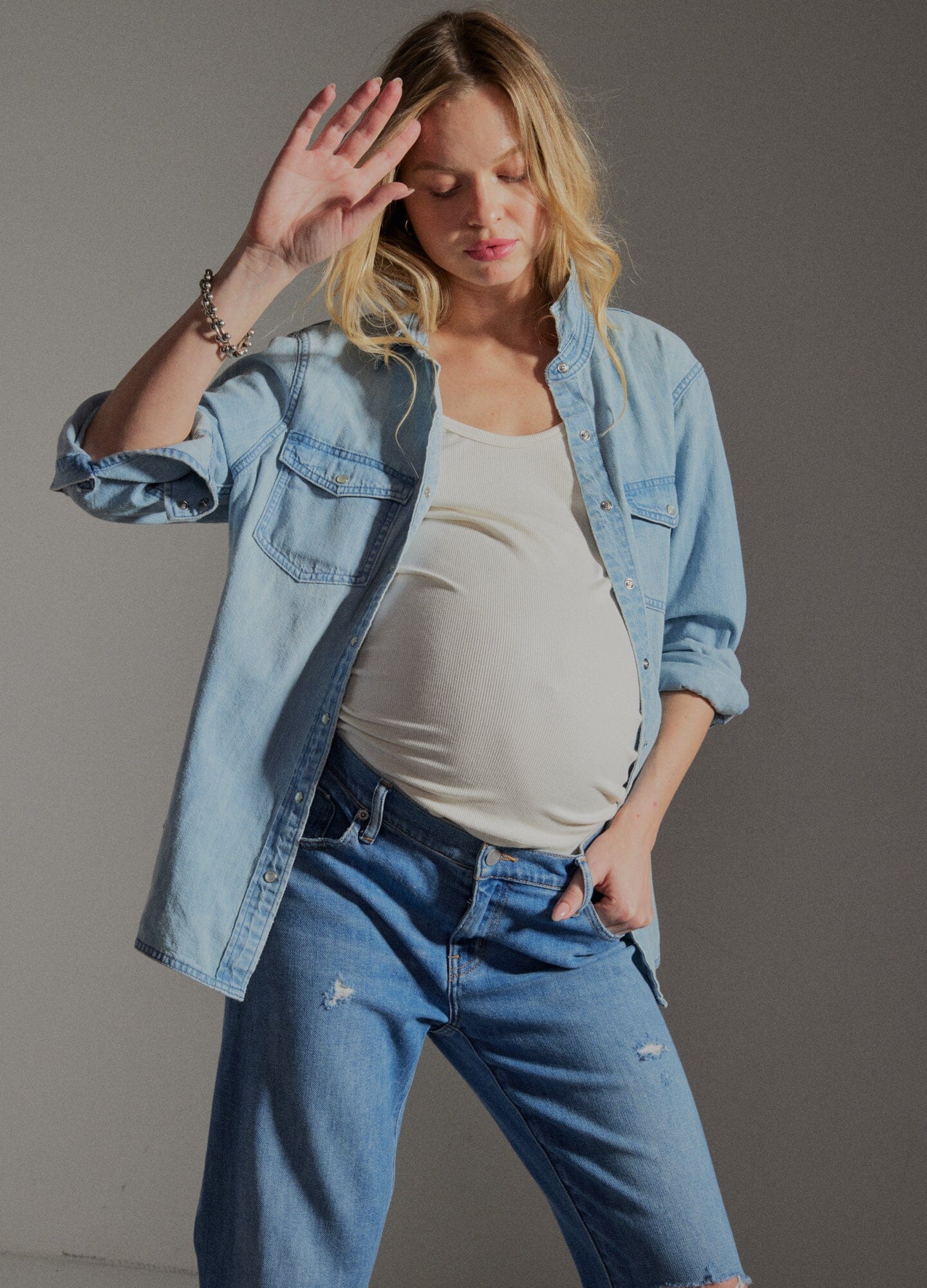 The Denim Maternity Shirt - Light wash denim maternity shirt with rolled sleeves, worn open over a white tank top during pregnancy.