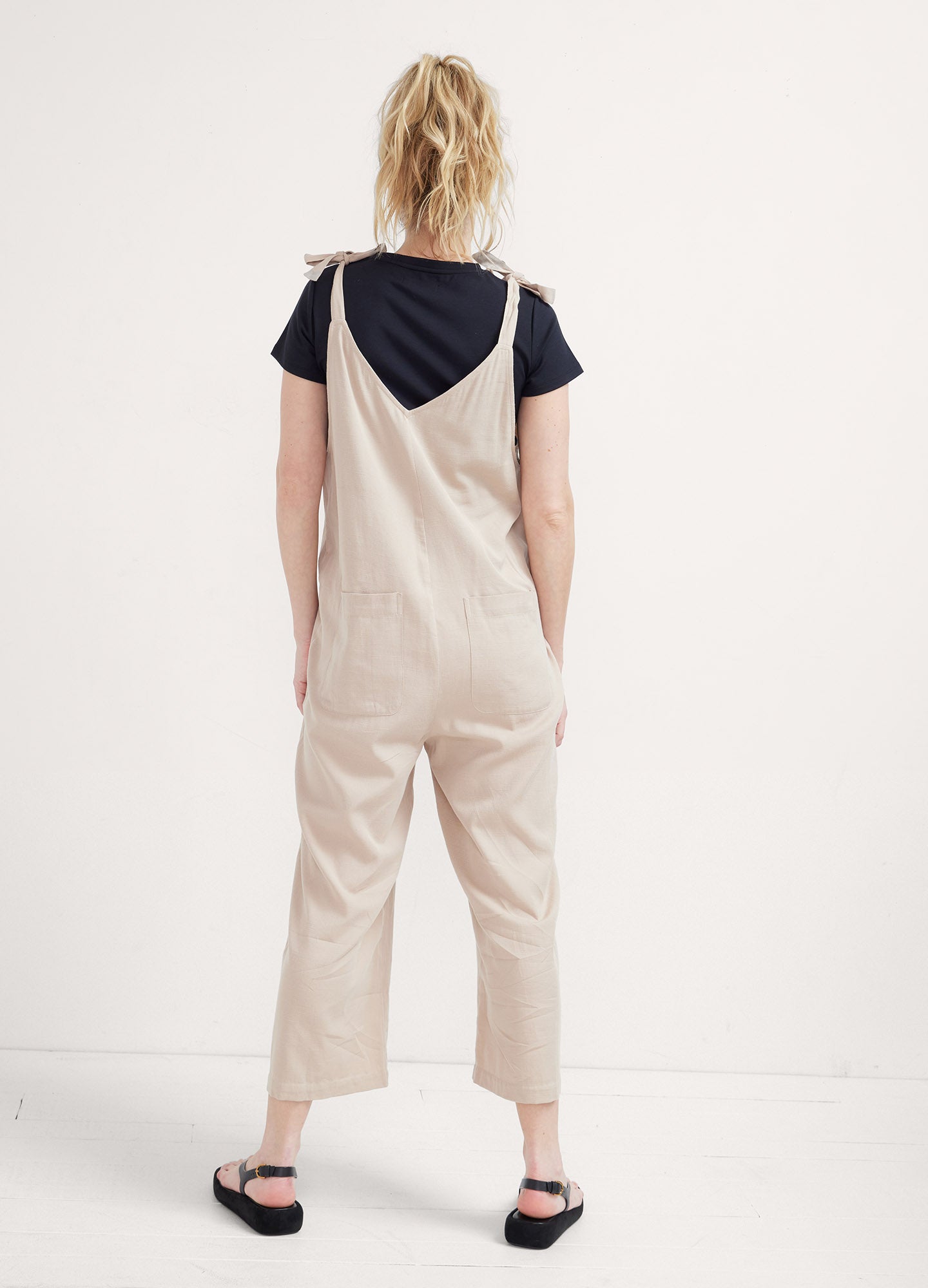 Light beige maternity jumpsuit with adjustable shoulder straps, loose fit, and cropped legs worn over a black short-sleeve.