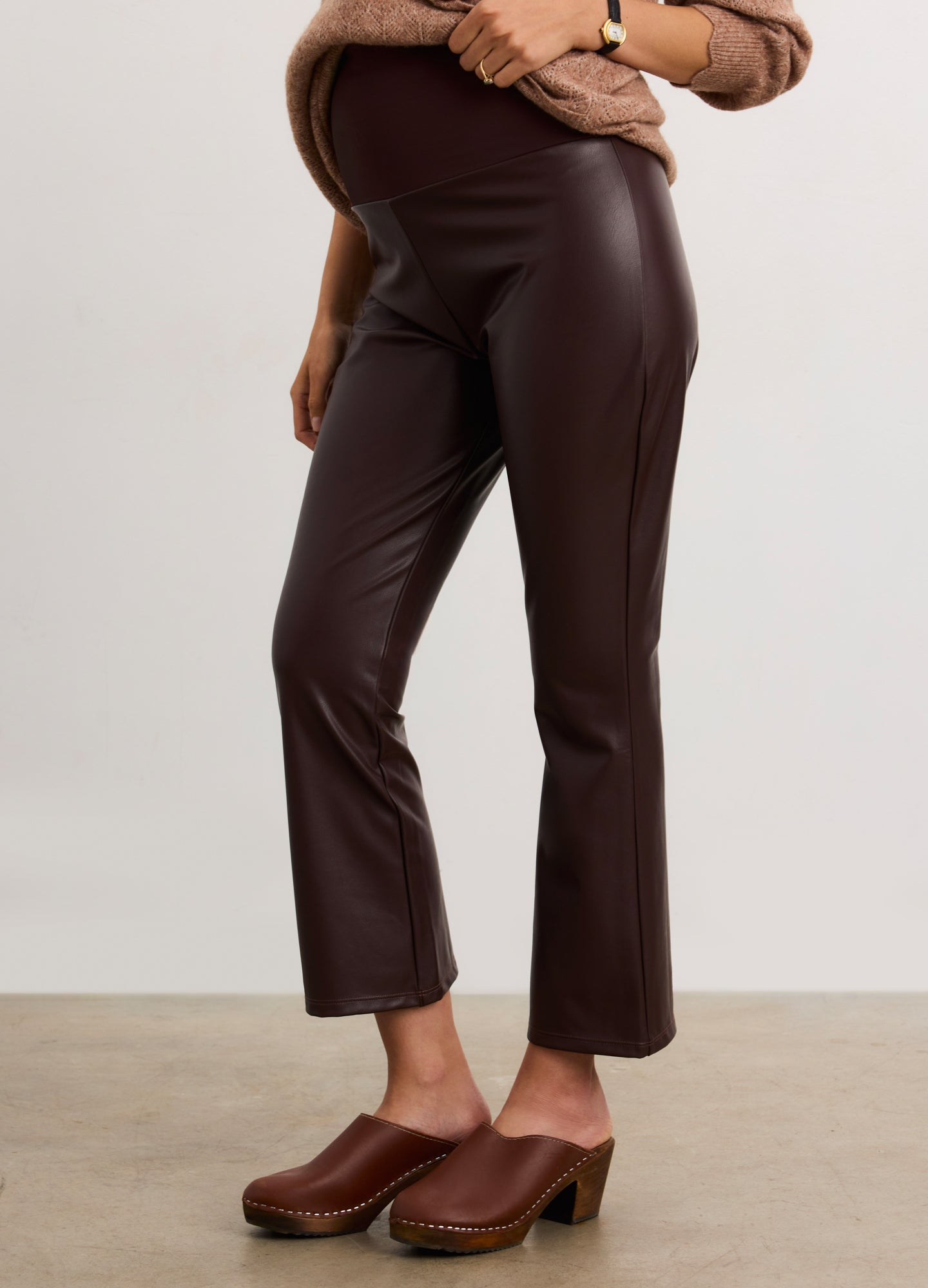 Pregnant woman wearing a brown sweater and leather pants against a white background