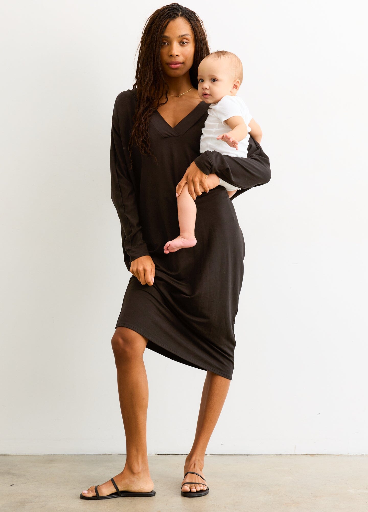 The Visitor Dress - Black maternity dress with long sleeves and V-neck, designed for pregnancy, nursing, and postpartum, modeled with baby.