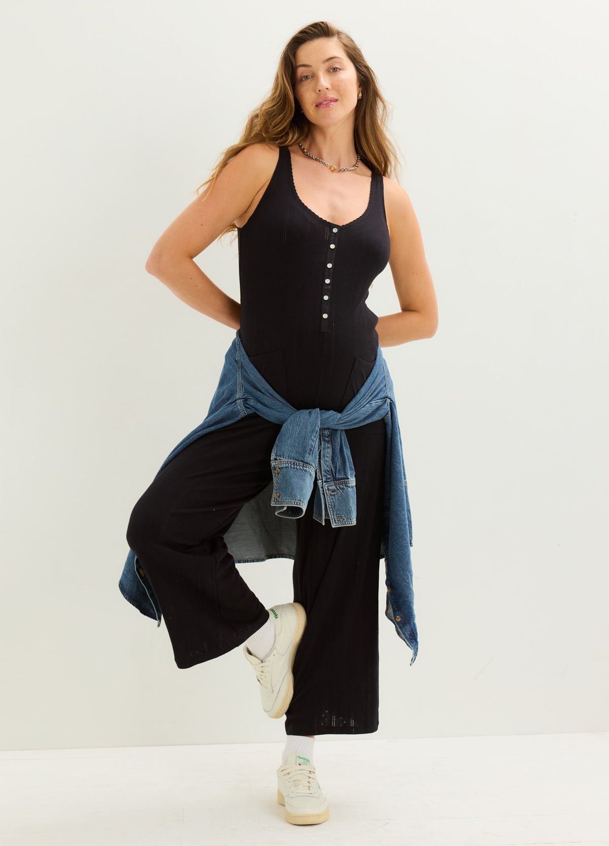 Black sleeveless maternity jumpsuit with buttoned front and wide-leg pants, styled with a tied denim jacket and white.