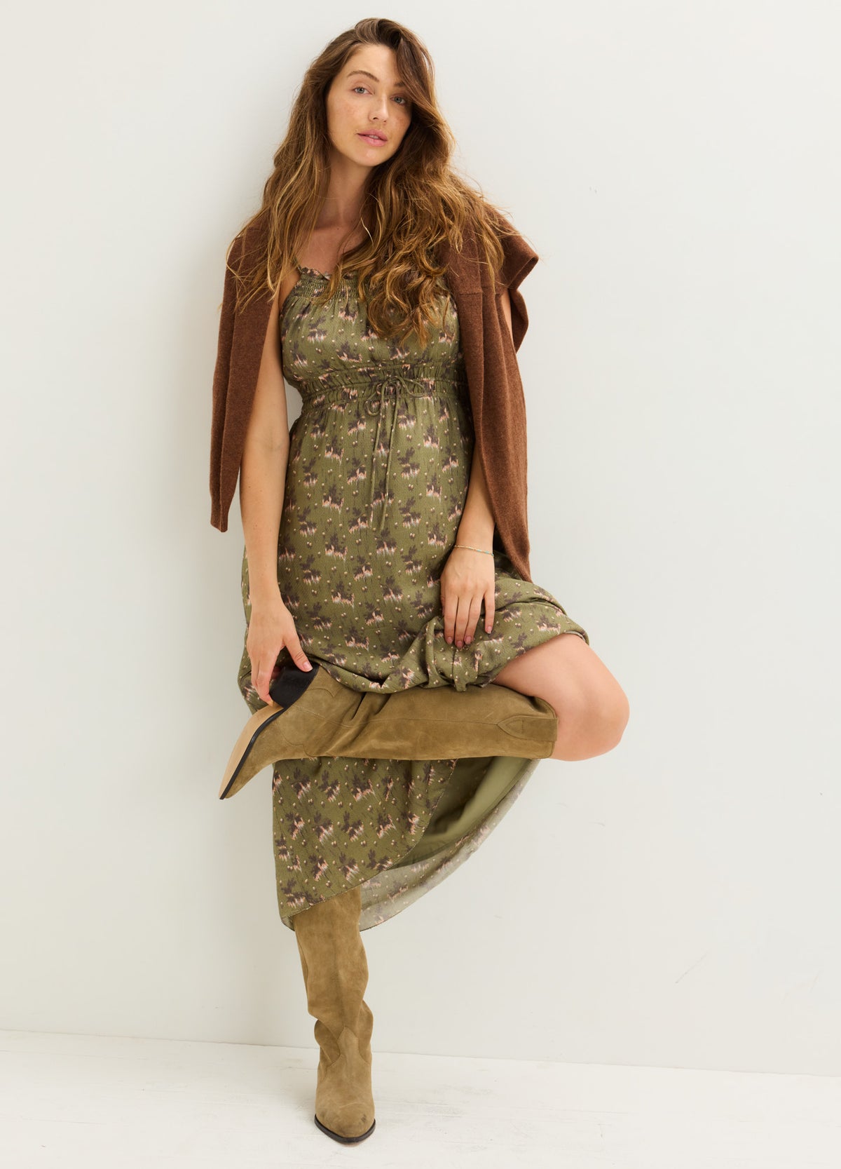 Olive green patterned maternity dress with sleeveless design, asymmetrical hem, paired with tan boots and brown cardigan.