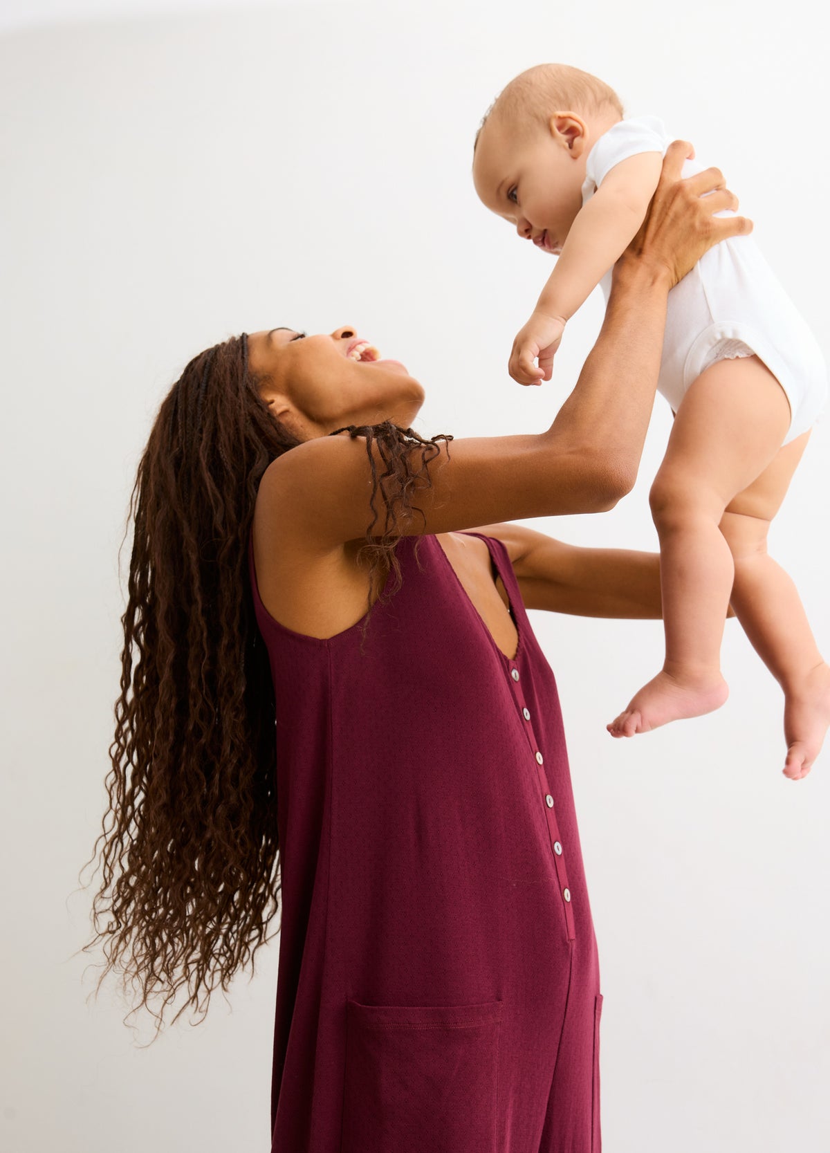 Sleeveless port-colored maternity jumpsuit with front buttons and pockets, worn by pregnant woman holding baby.