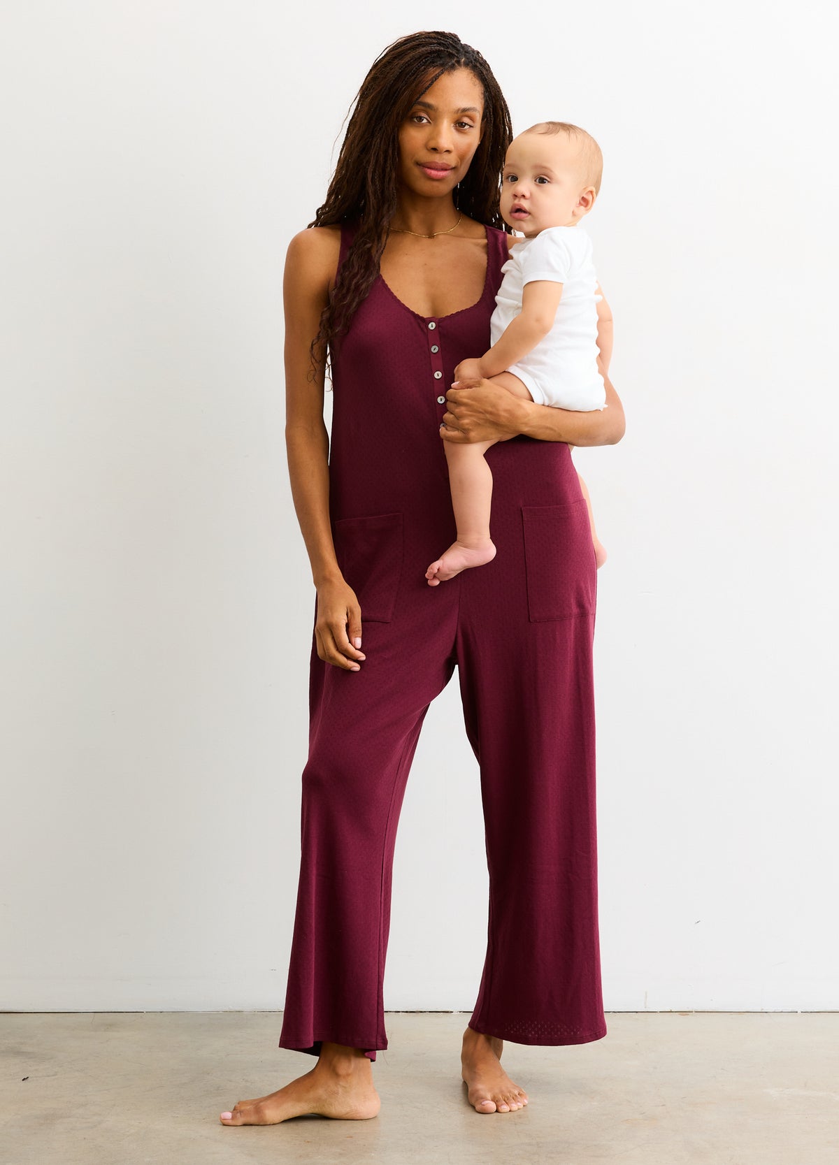 Sleeveless port-colored maternity jumpsuit with wide legs, modeled by pregnant woman holding baby.