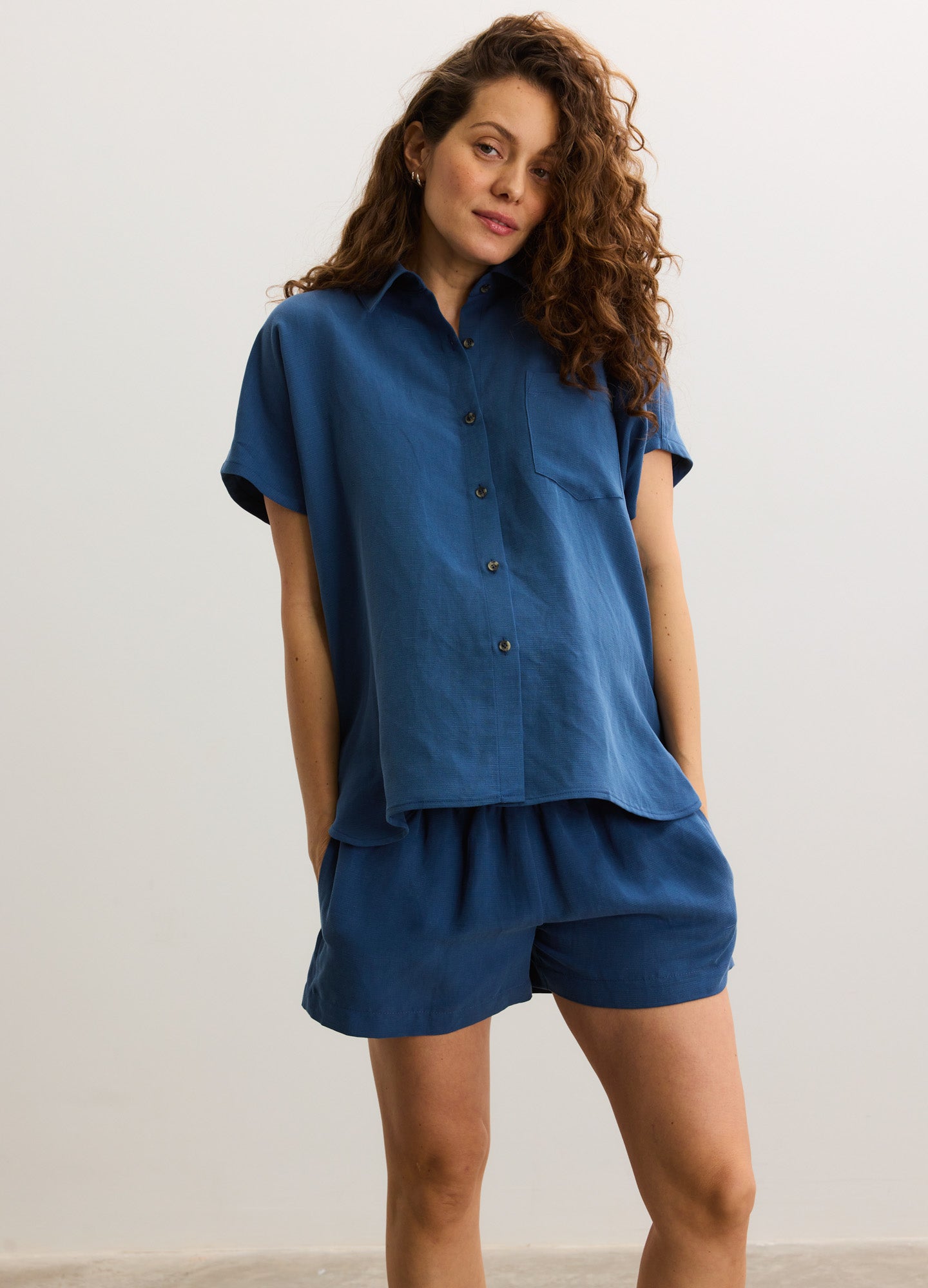 Loose-fitting maternity shorts in deep blue with an elastic waistband, paired with a matching short-sleeve button-up shirt.