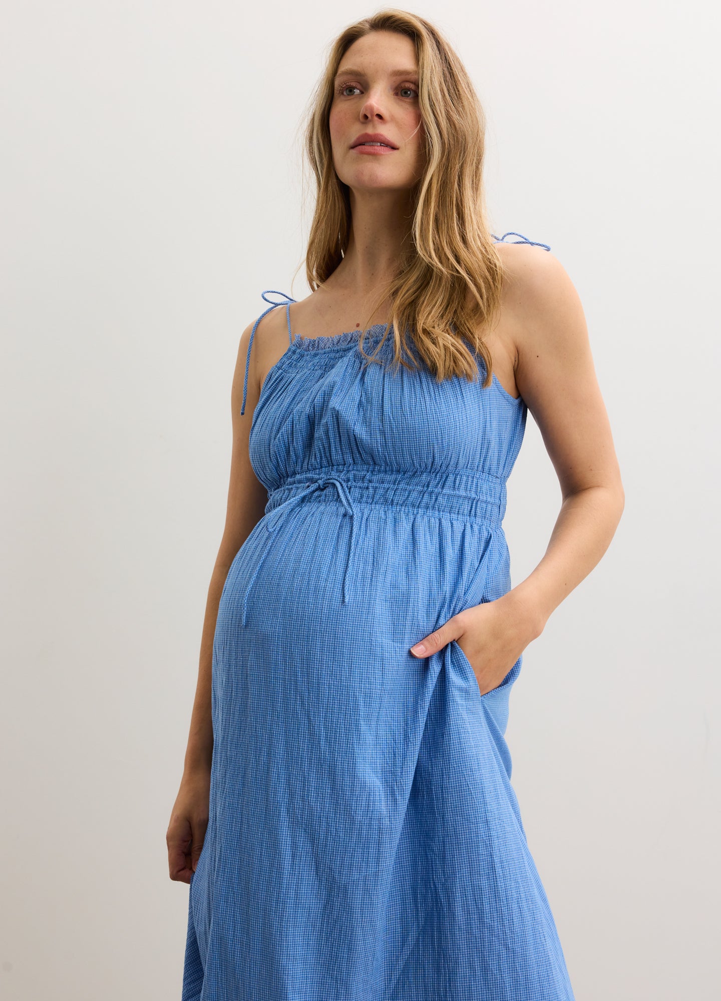 Blue sleeveless maternity dress with adjustable tie straps and gathered bodice, cinched waist with side pockets.