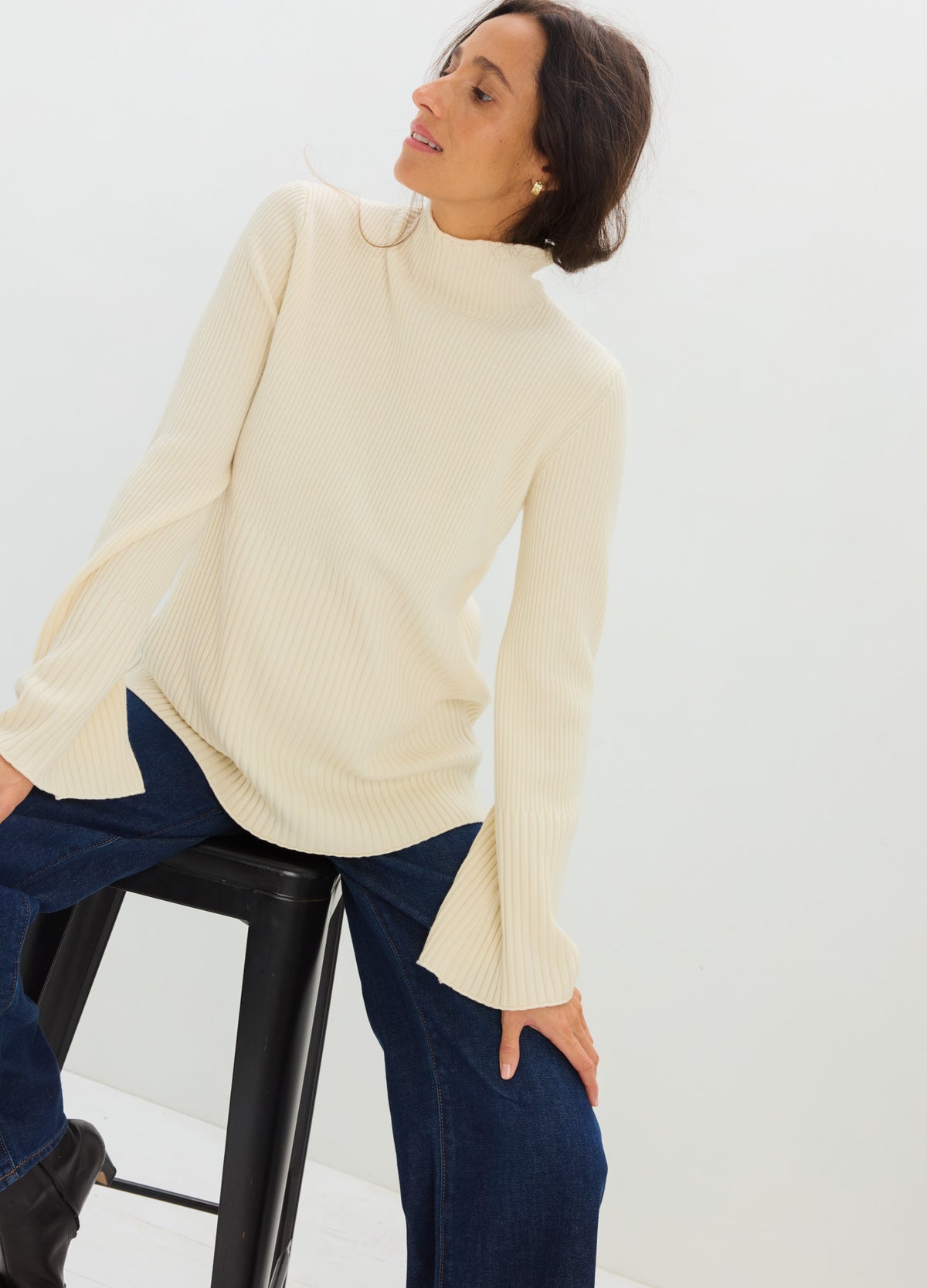 Cream-colored knit maternity turtleneck sweater with long, flared sleeves and relaxed fit over dark jeans.