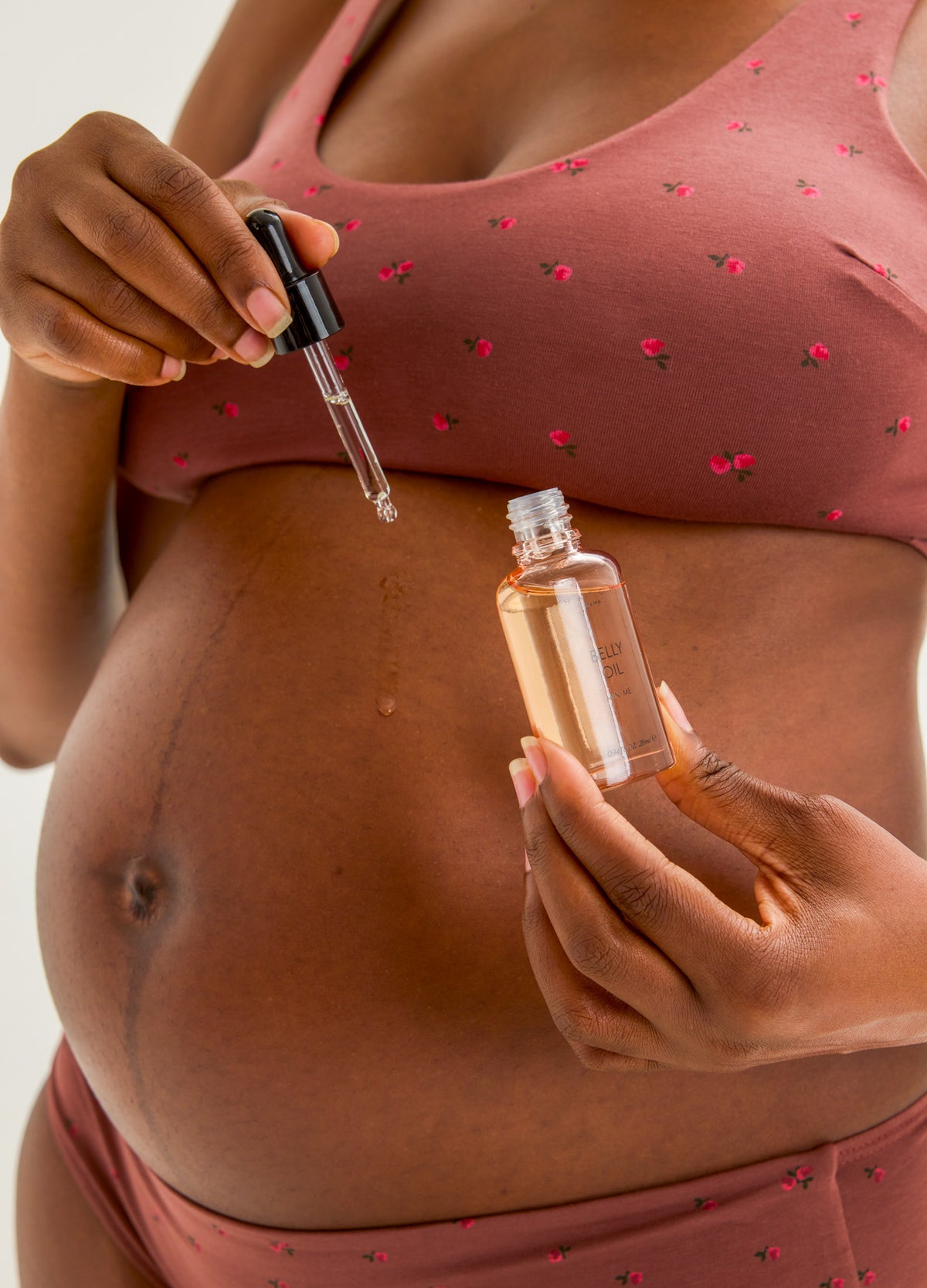 Clear glass bottle of light amber belly oil with dropper held by pregnant woman in pink maternity bra with small red flower.