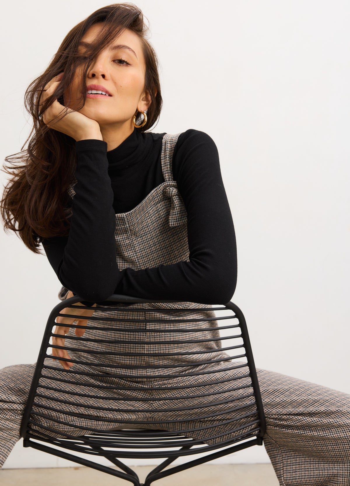 Pregnant woman wearing mini houndstooth maternity soft stretch onesie over black long sleeve top sitting behind black wire.