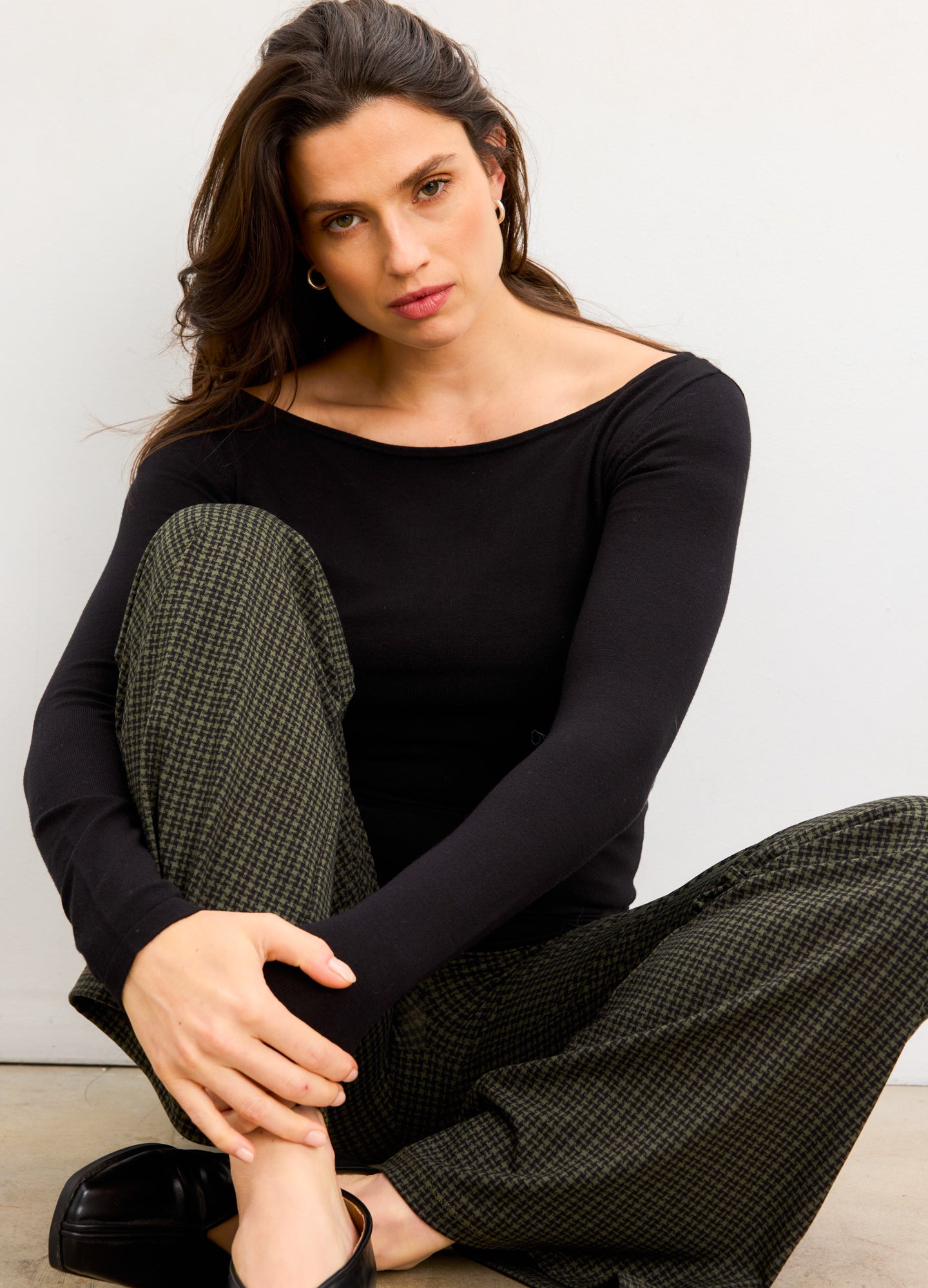 Pregnant woman wearing a black top and plaid pants sitting on a white background