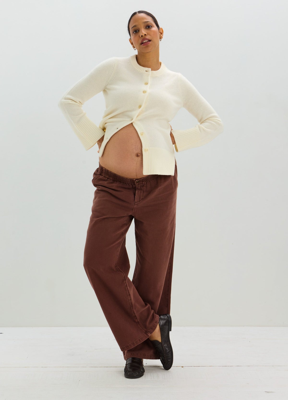 Pregnant model wearing a cream button-front cardigan with brown maternity trousers and black shoes.