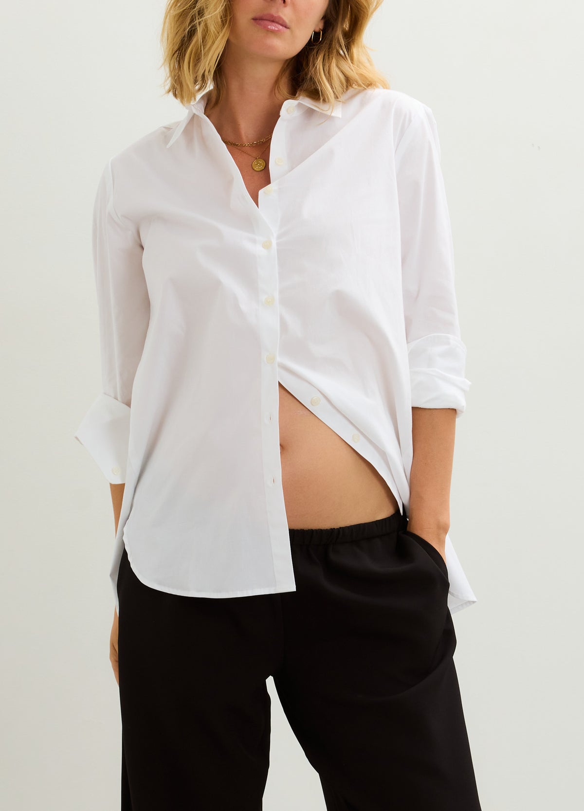 White long-sleeve maternity buttondown shirt with relaxed fit and curved hem, styled open over black pants.