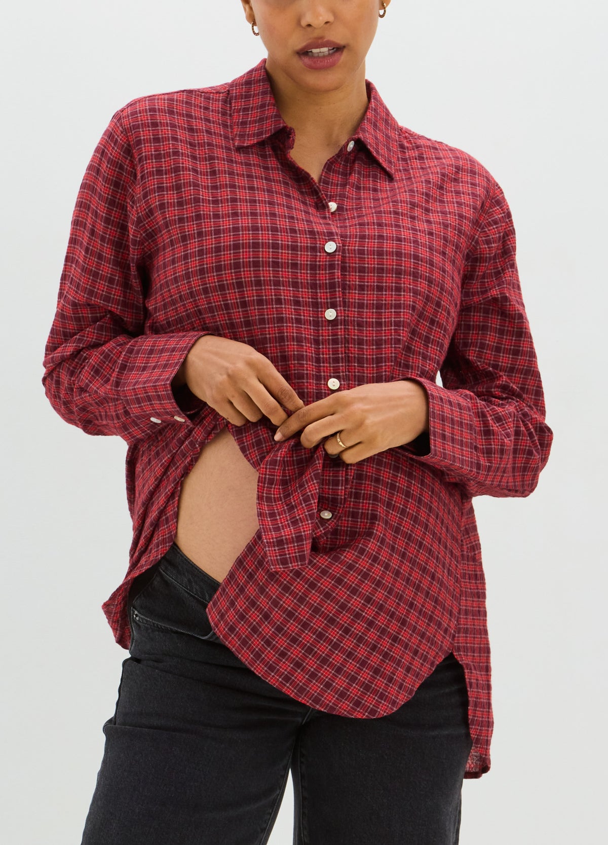 Red and black plaid maternity buttondown shirt with long sleeves and curved hem, worn open over black pants.