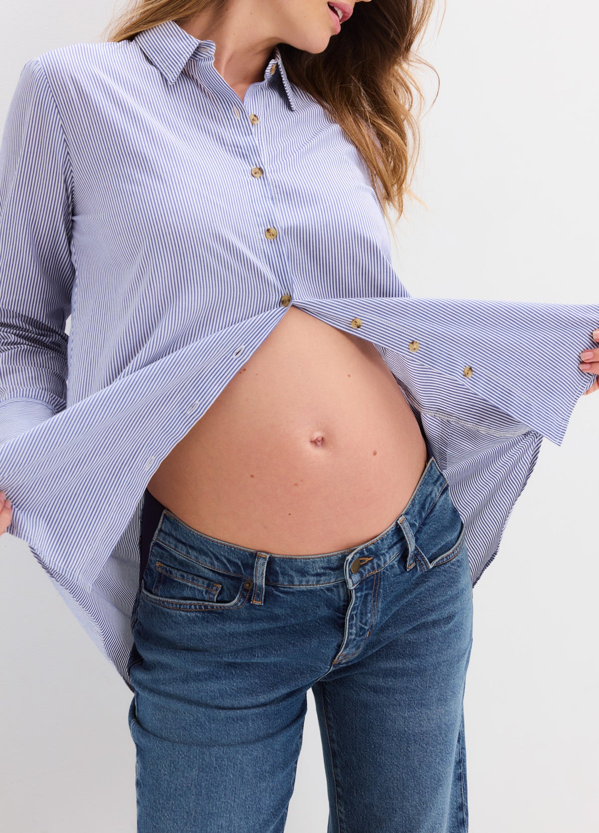 Pregnant woman wearing a light blue buttondown shirt open over her belly, paired with blue jeans.
