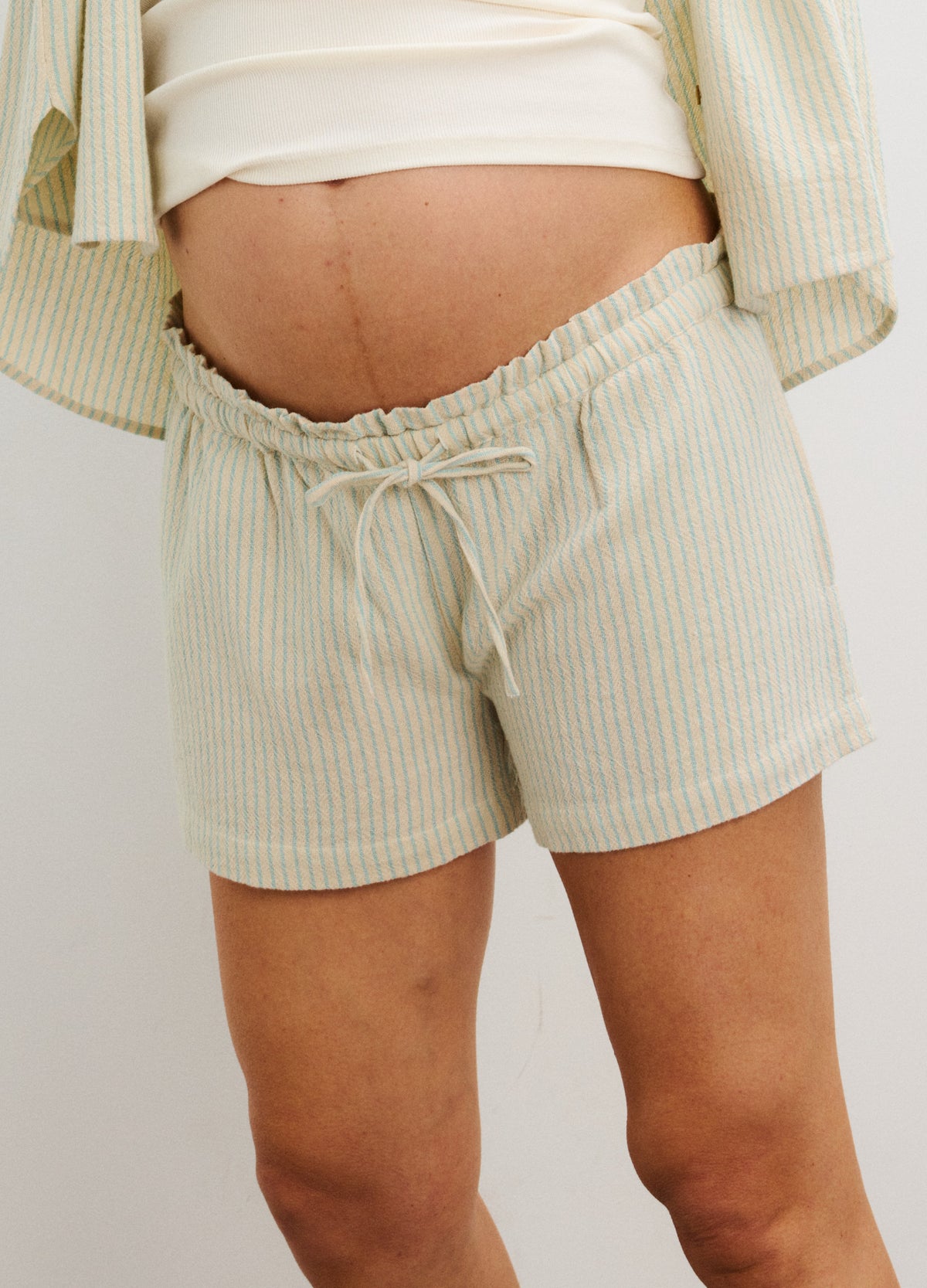 Light beige maternity beach shorts with thin white vertical stripes, elastic waistband, and drawstring tie.