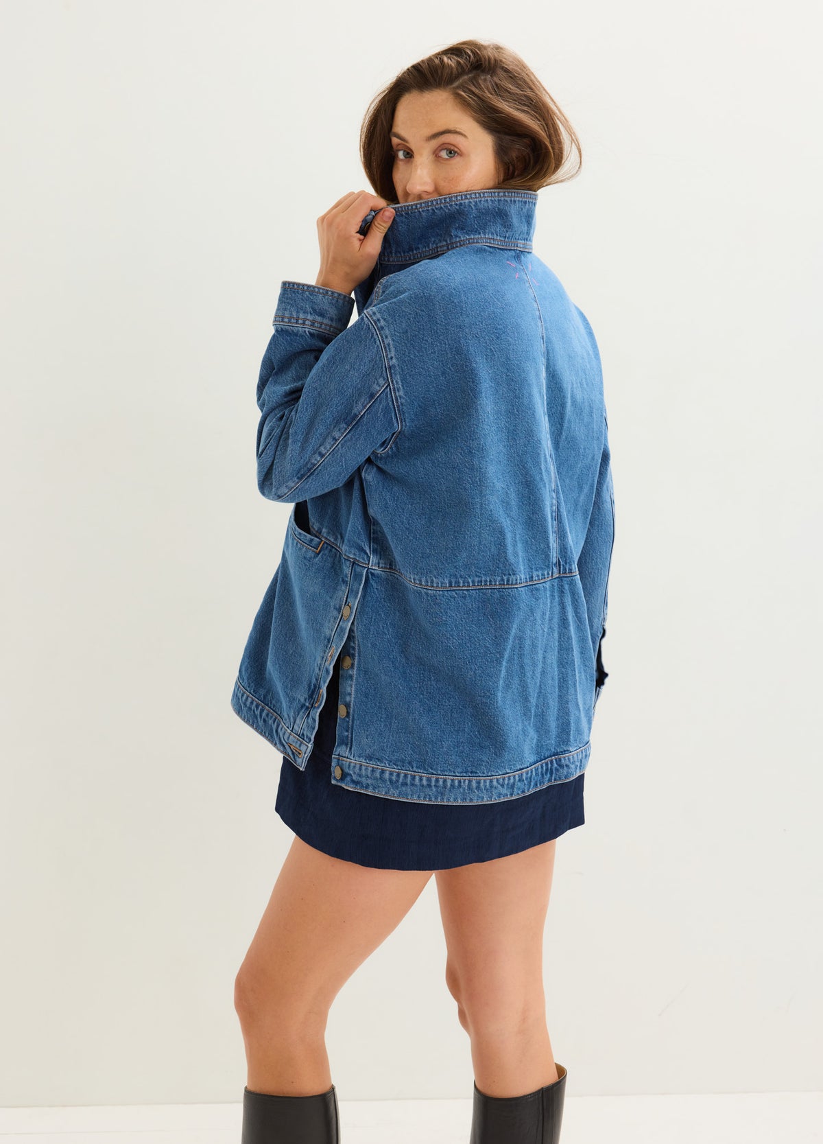 Blue denim maternity utility jacket with high collar and side button vents, worn over a dark dress.