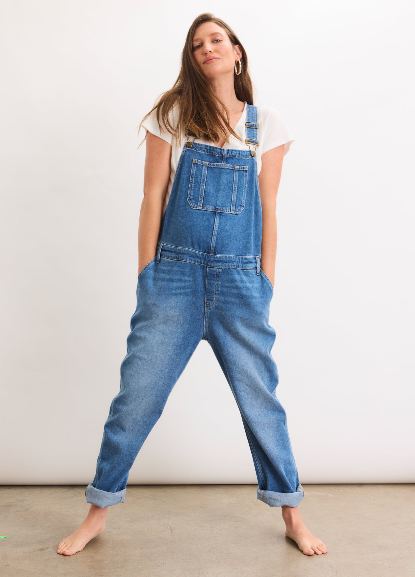 Blue denim maternity overall with adjustable straps and front pocket, worn over white short-sleeve top.