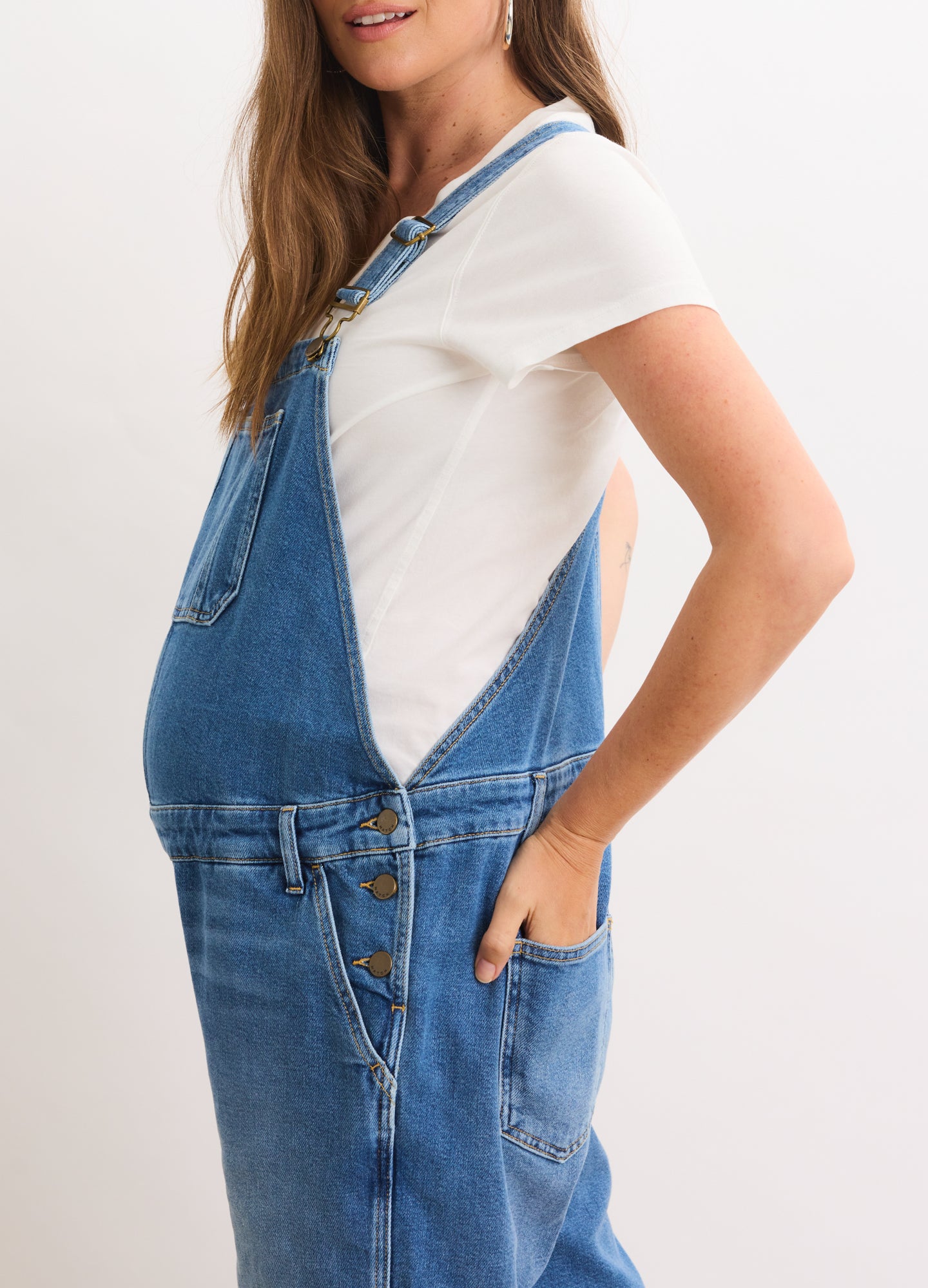 Blue denim maternity overall with adjustable straps and side buttons, worn over a white short-sleeve shirt.