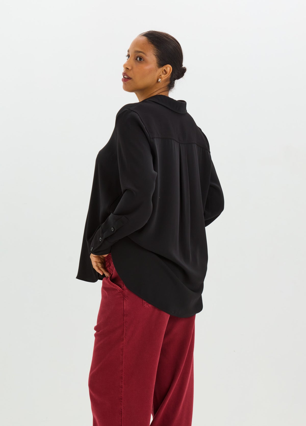 Woman wearing a relaxed black drapey button-down shirt with long sleeves and a curved hem over red trousers.