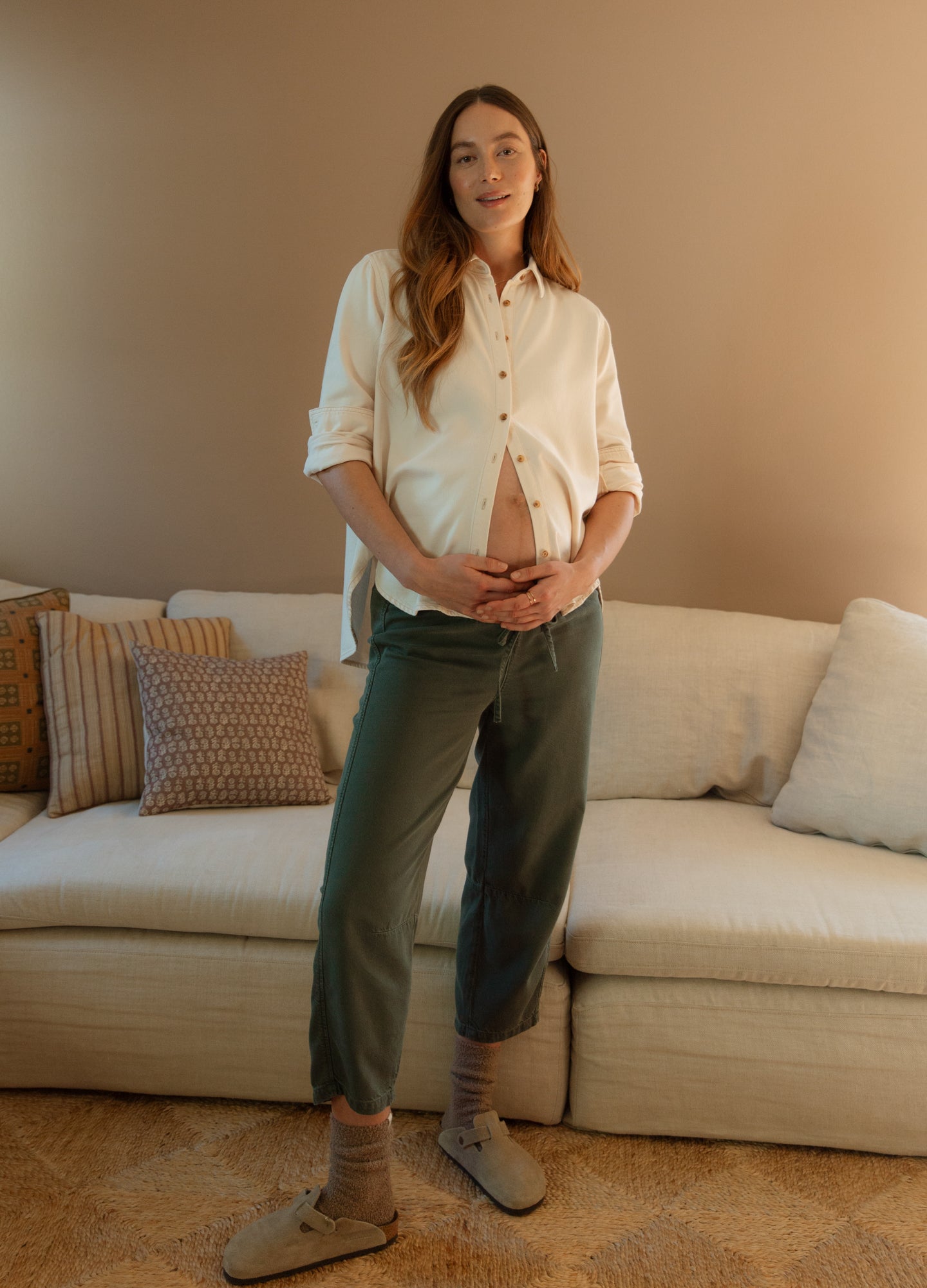Maternity dark olive drapey denim barrel pants with relaxed fit and cuffed hems worn with a cream button-up shirt and tan.