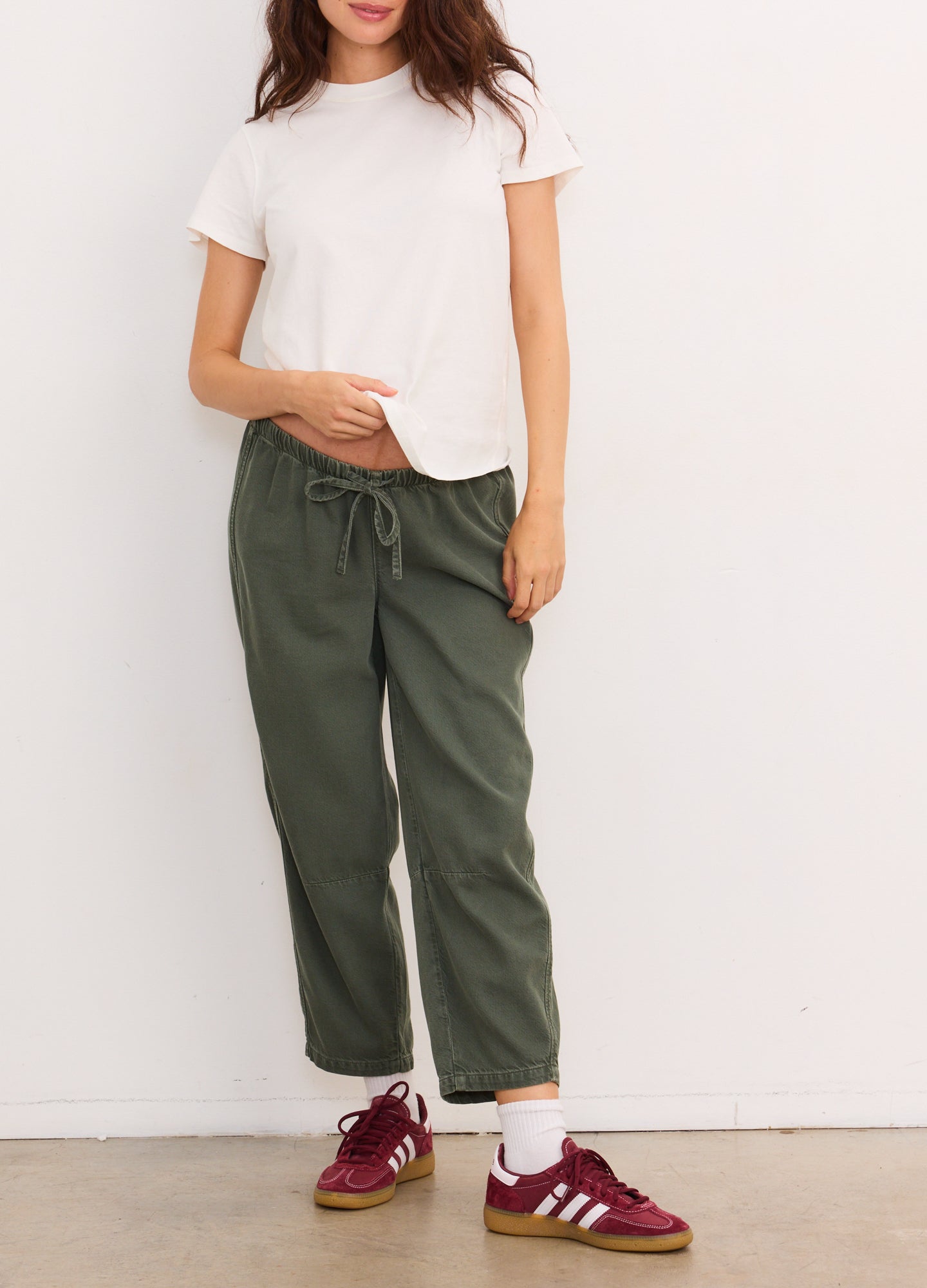 Olive green drapey denim maternity barrel pants with elastic drawstring waist, styled with white tee and burgundy sneakers.