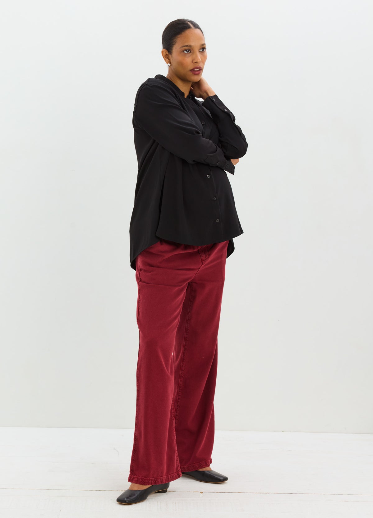 Pregnant woman wearing burgundy drapey maternity denim pants with a loose black blouse and black shoes.