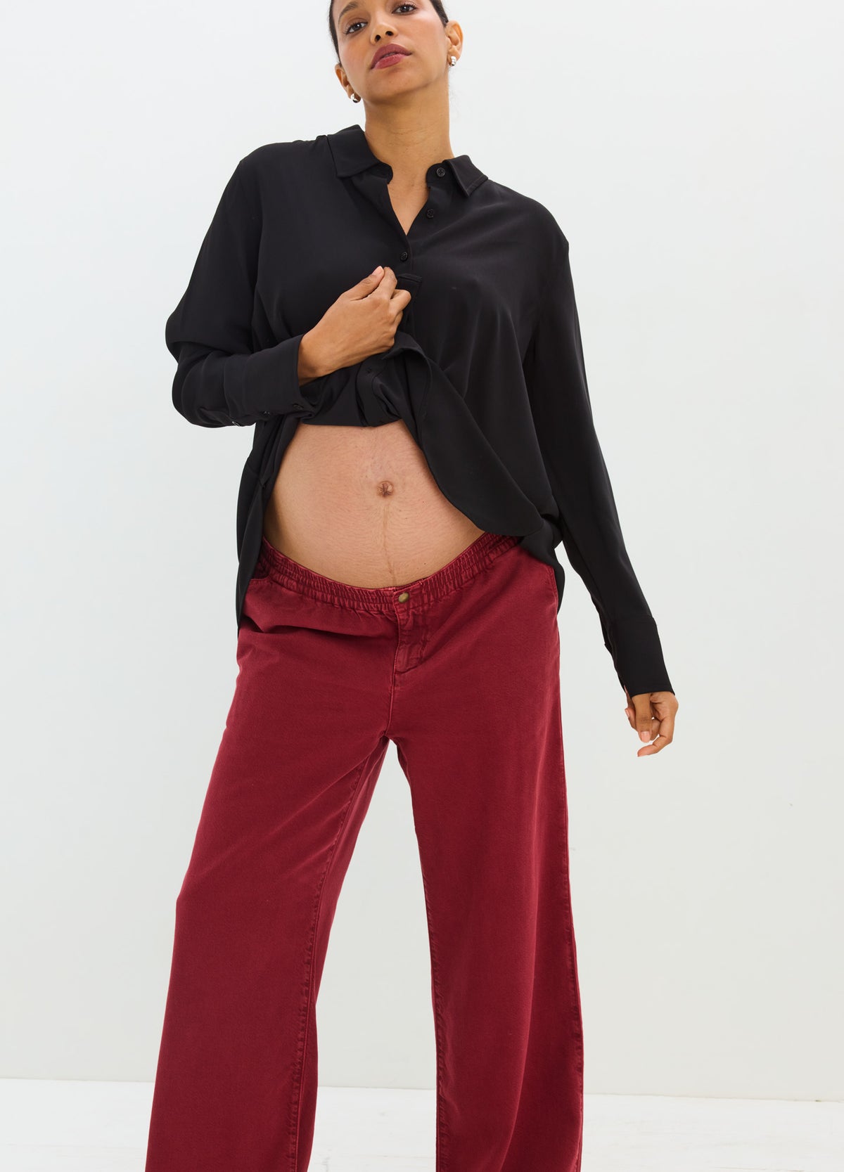 Maternity drapey denim pants in deep red with relaxed fit and elastic waistband, modeled with black button-up shirt.