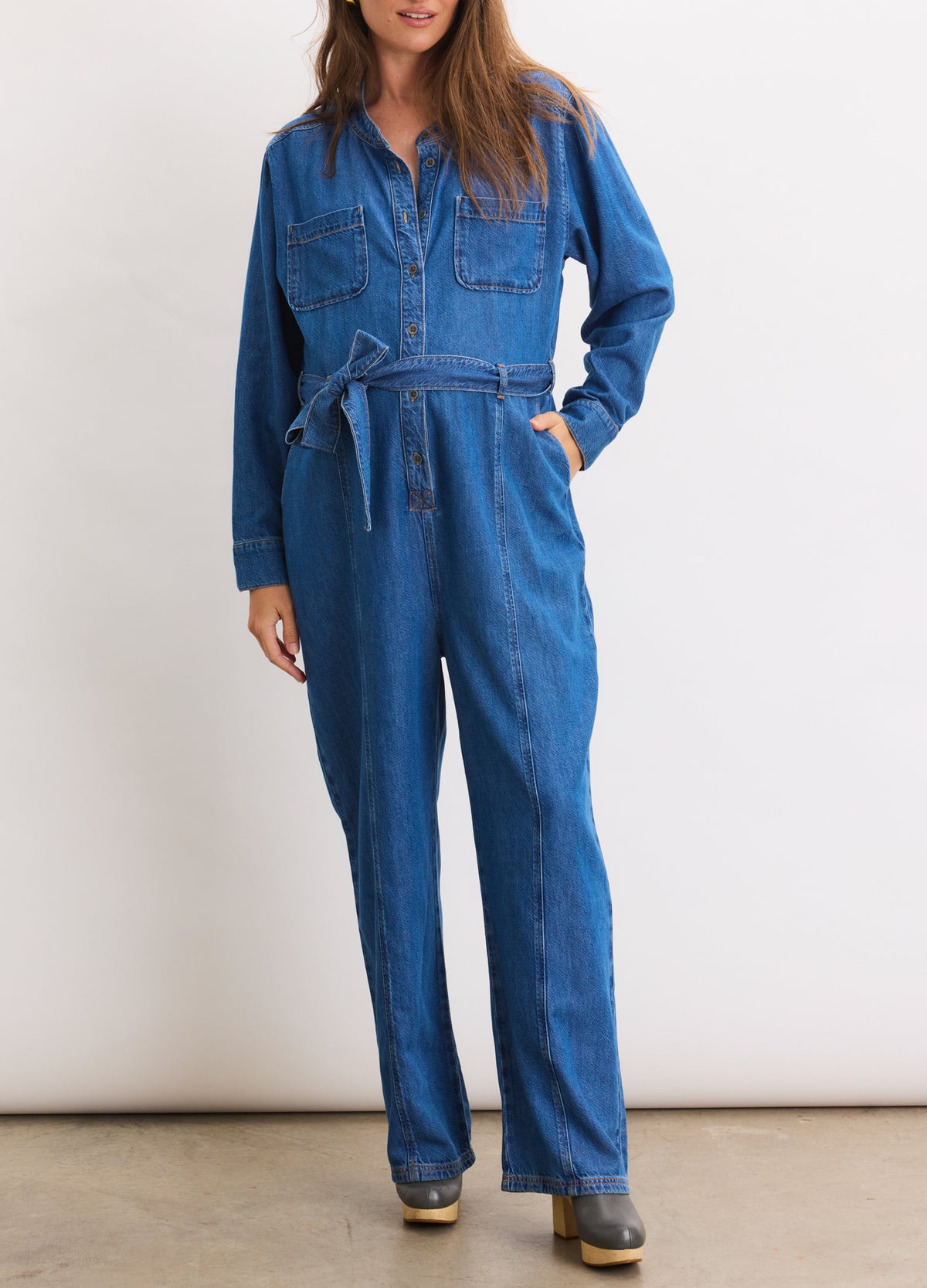 Blue denim jumpsuit with long sleeves, button-up front, and waist tie, designed for maternity.
