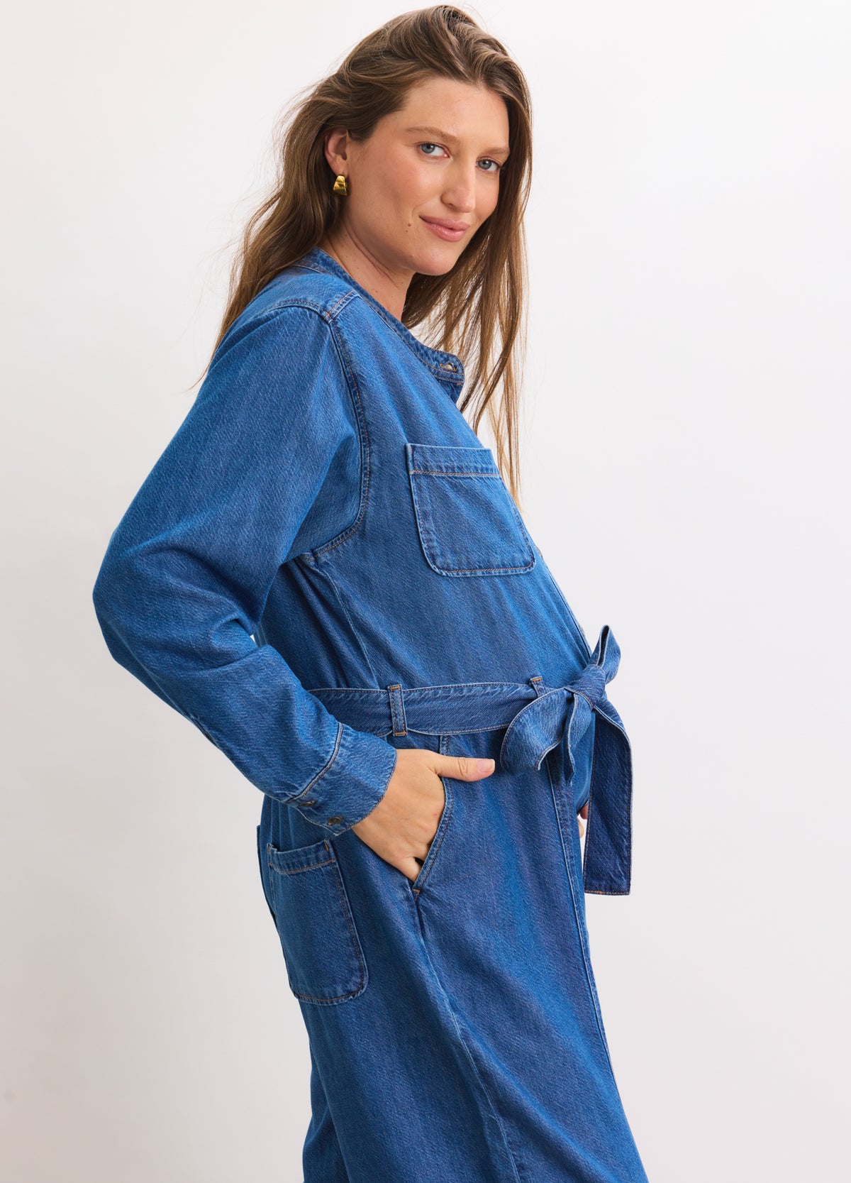 Blue denim maternity jumpsuit with long sleeves, front chest pocket, side pockets, and a tie waist belt.