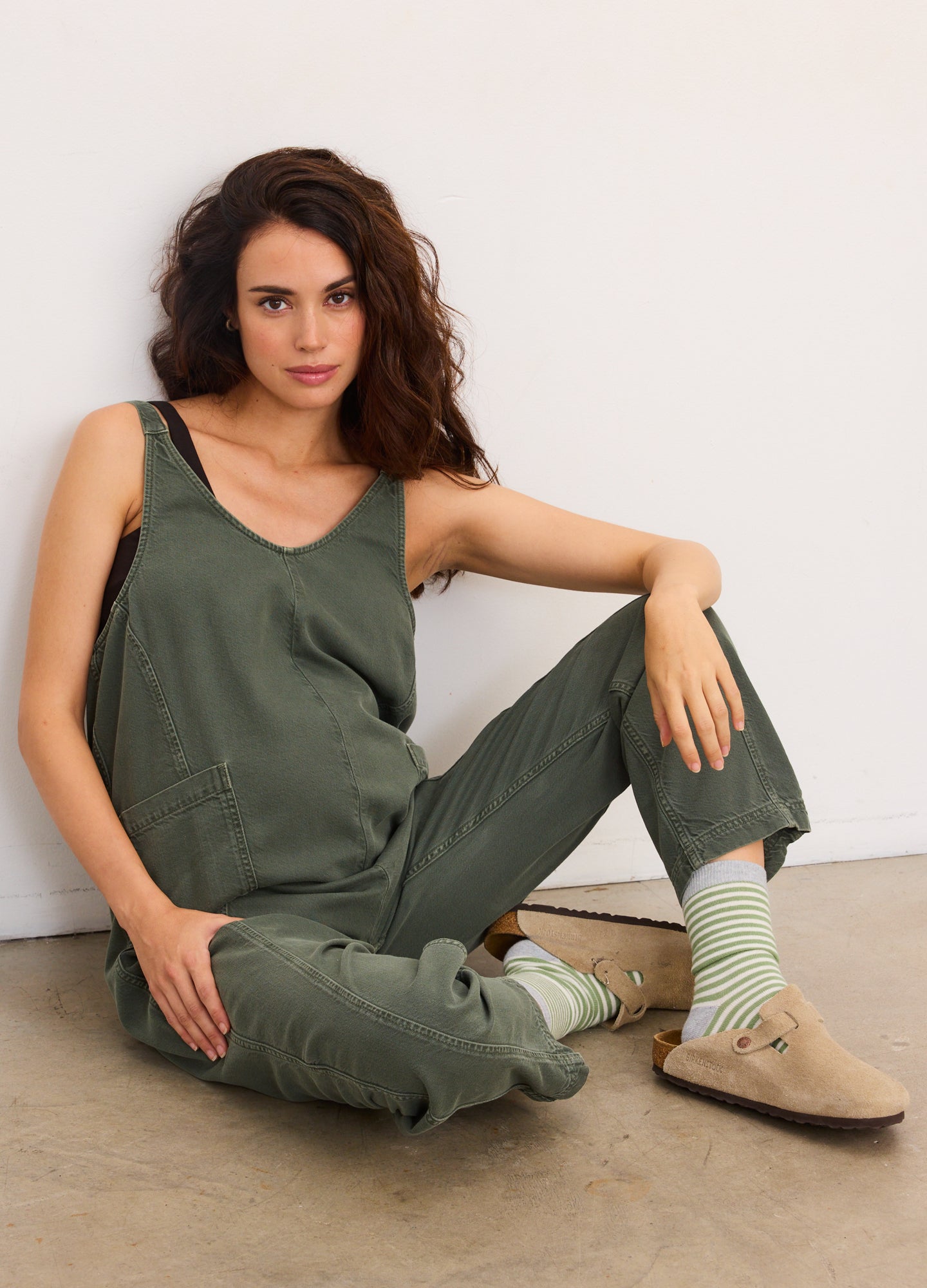 Olive green sleeveless maternity jumpsuit with adjustable straps and relaxed fit, worn with striped socks and beige slip-on.