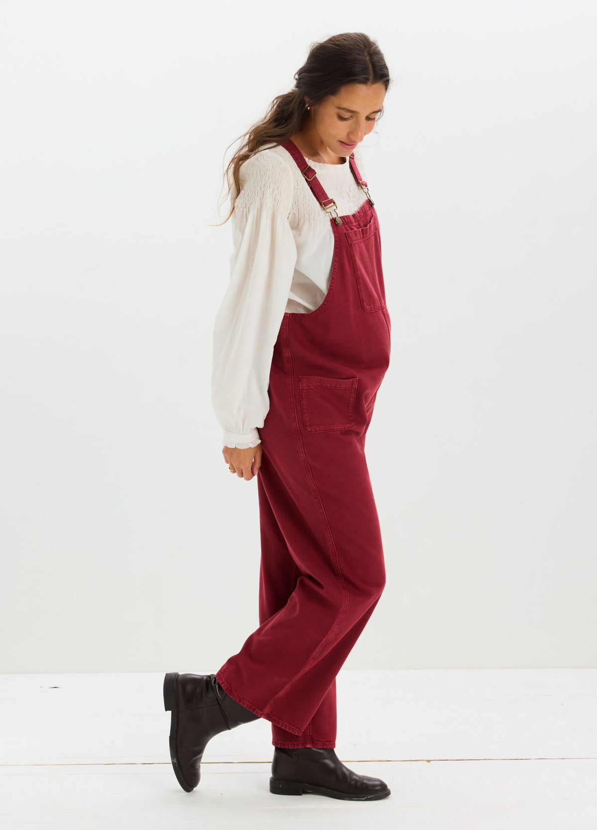 Red maternity denim overalls with adjustable straps, loose fit, styled with a white long-sleeve top and black boots.