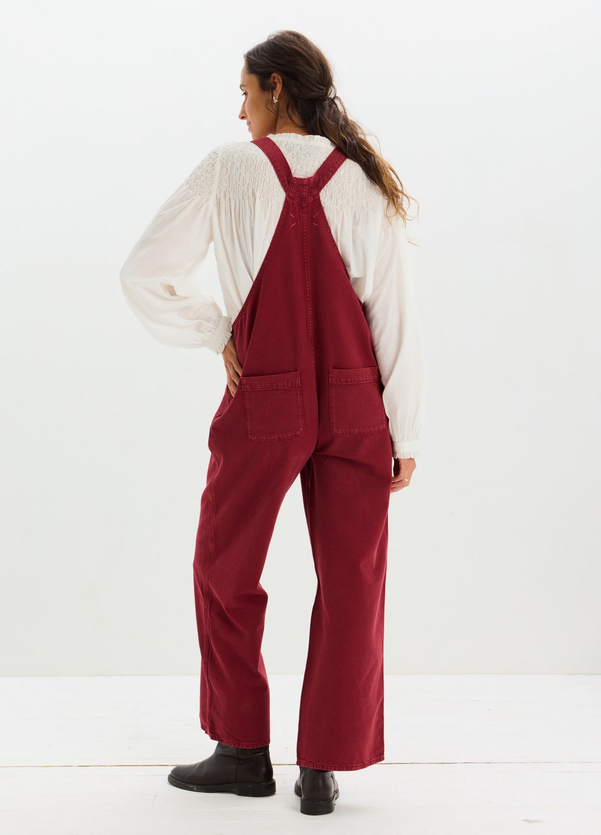 Maternity drapey denim overalls in deep red with wide legs and adjustable shoulder straps worn over a white long-sleeve top.