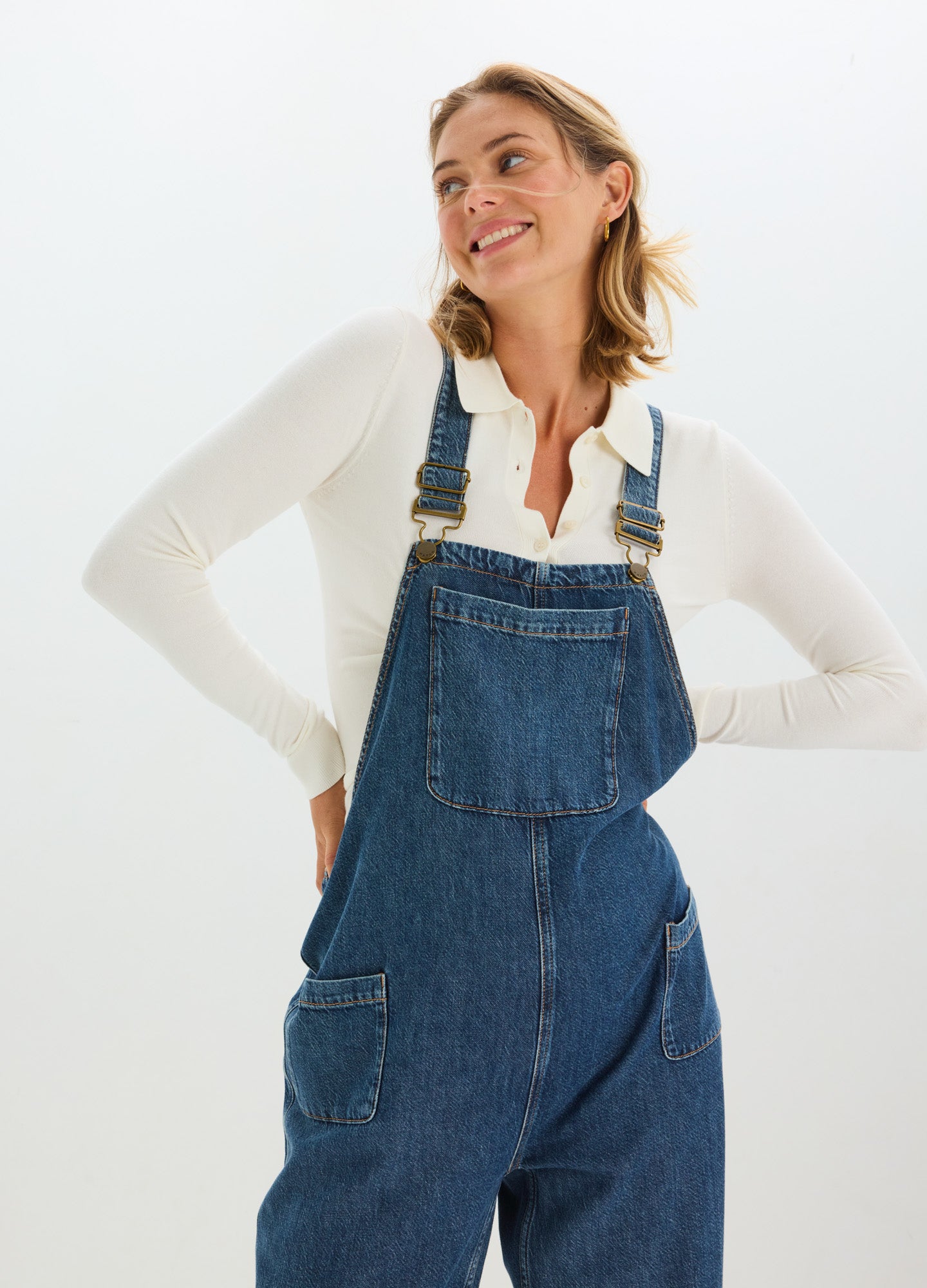Blue denim maternity overalls with adjustable shoulder straps and large front pocket worn over a white long-sleeve top.