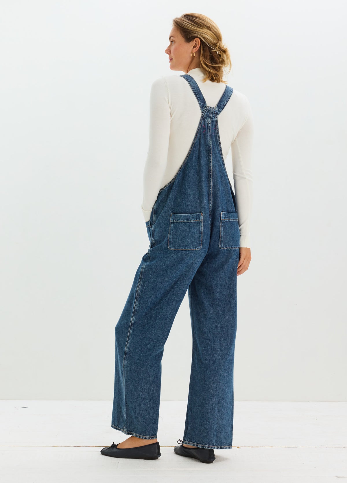 Dark blue denim maternity overalls with adjustable cross-back straps and wide-leg fit, worn over a white long-sleeve top.