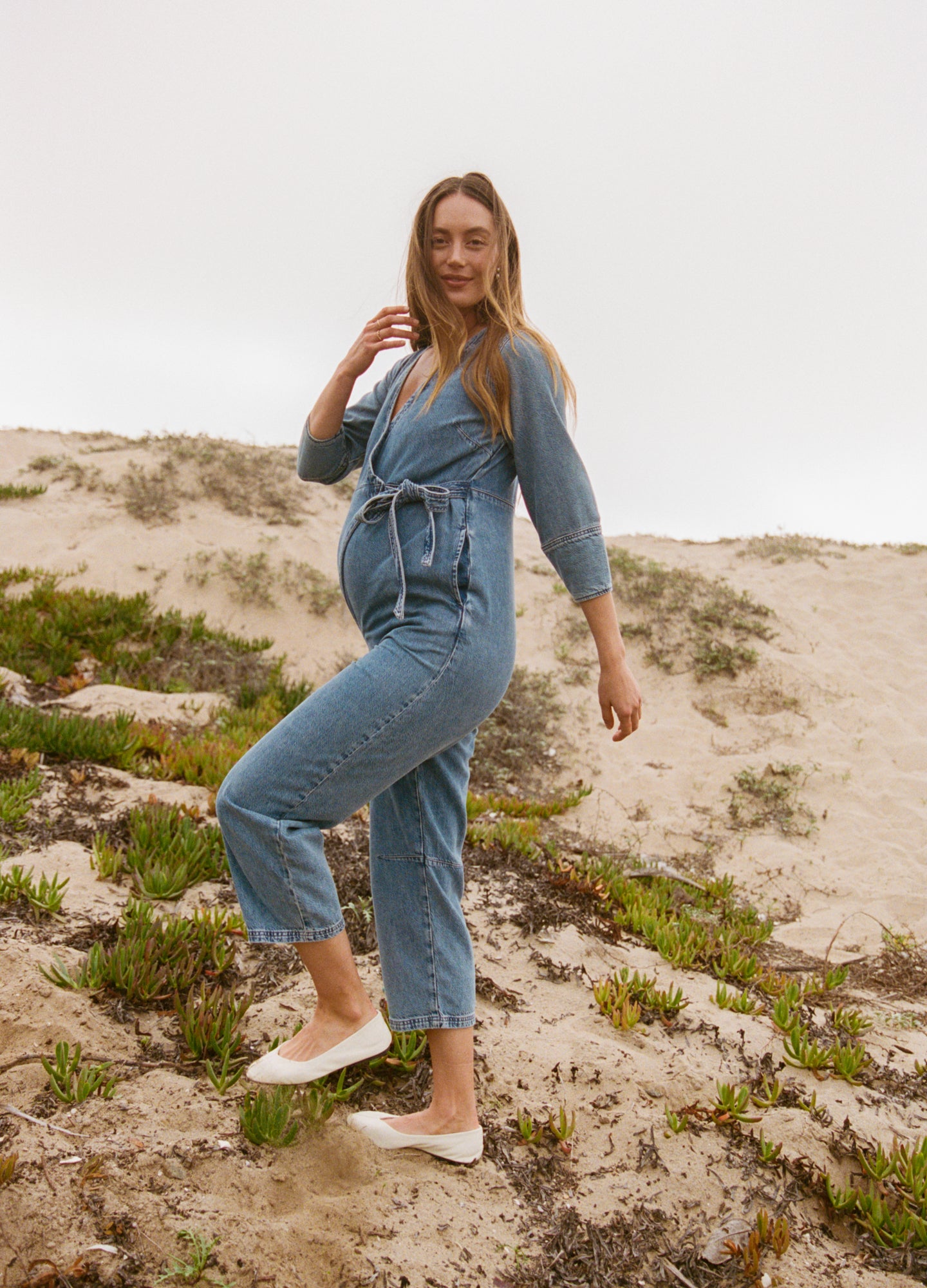 Light blue maternity denim jumpsuit with long sleeves, front buttons, and a tie waist belt.