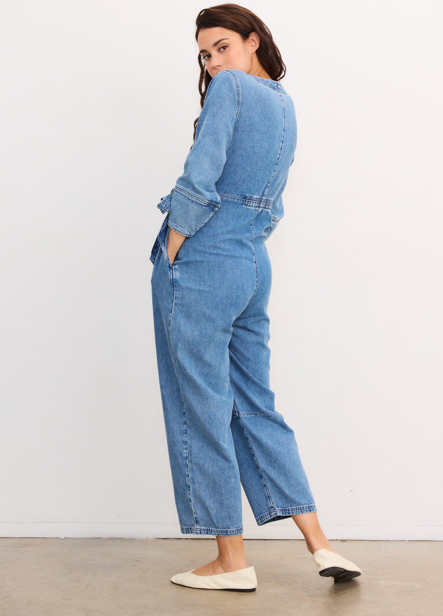 Light blue denim maternity jumpsuit with long sleeves, cinched waist, and wide legs, paired with white slip-on shoes.