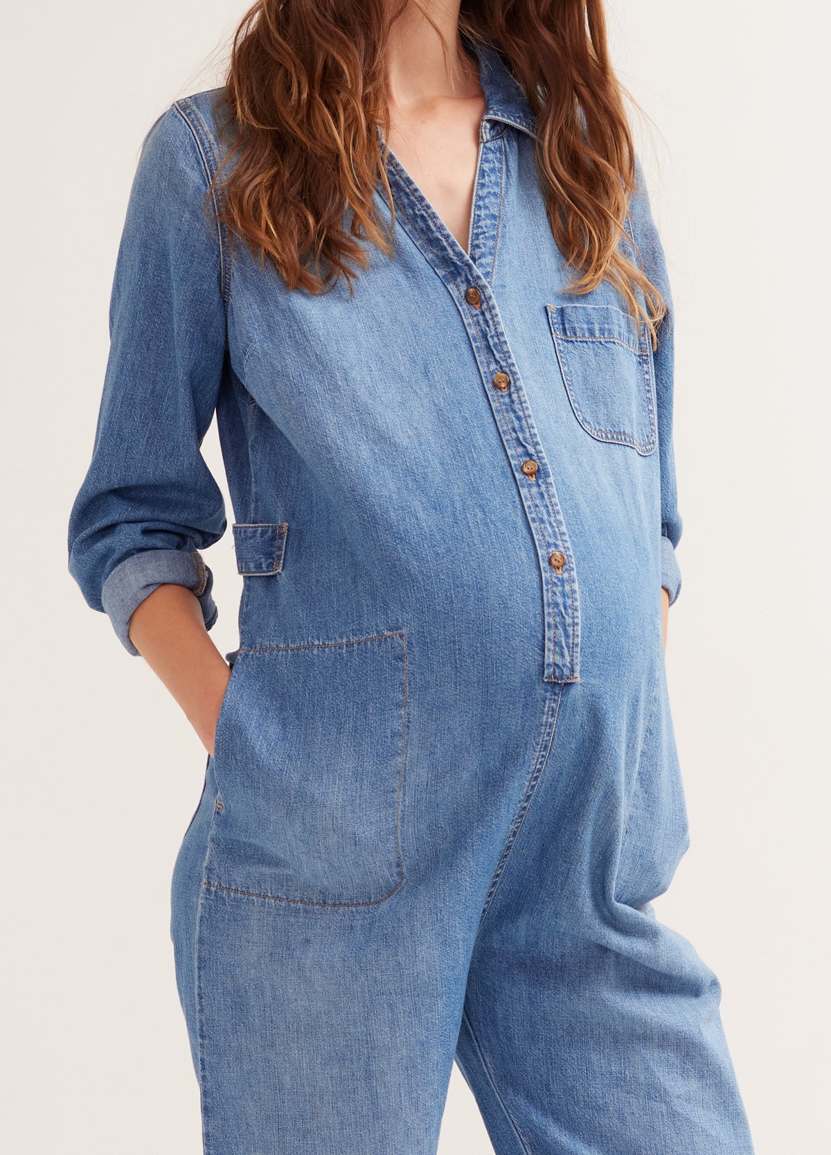 Indigo denim maternity jumpsuit with long sleeves, button front, and side pockets on pregnant model.