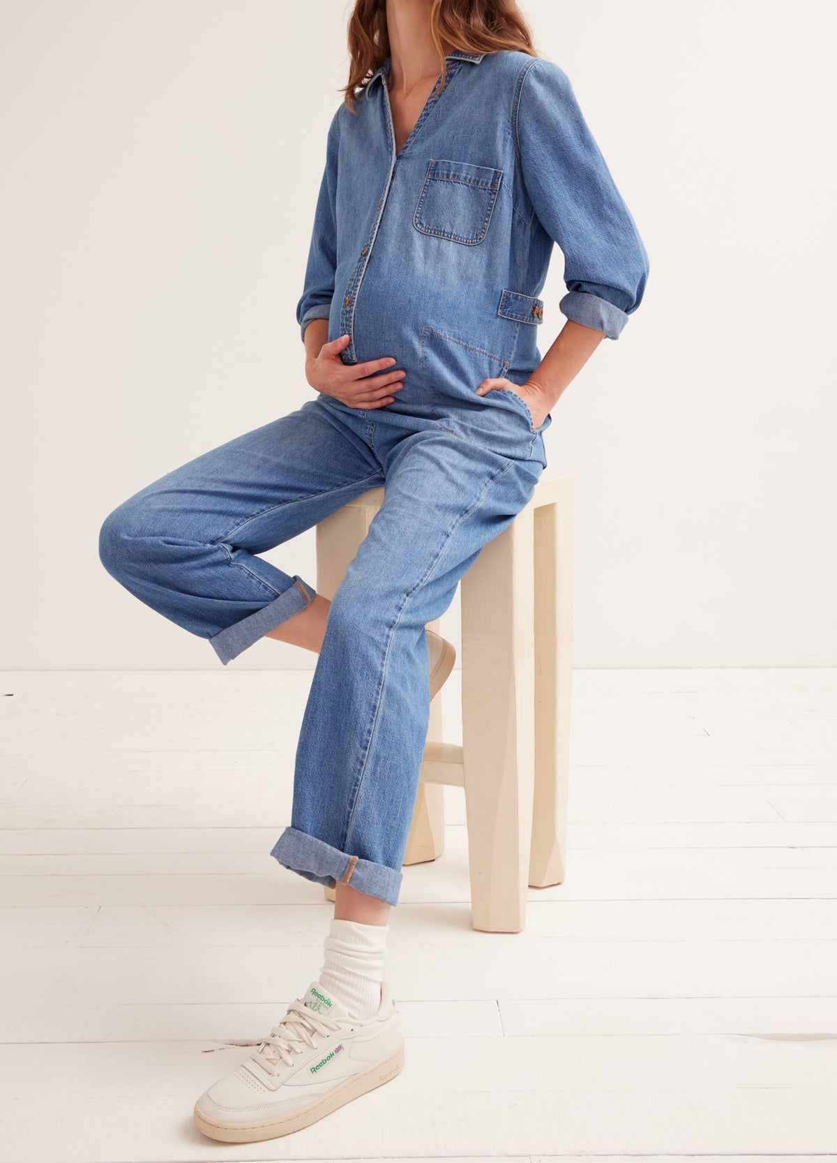 Pregnant woman wearing indigo maternity denim jumpsuit with zip front and rolled cuffs sitting on stool.