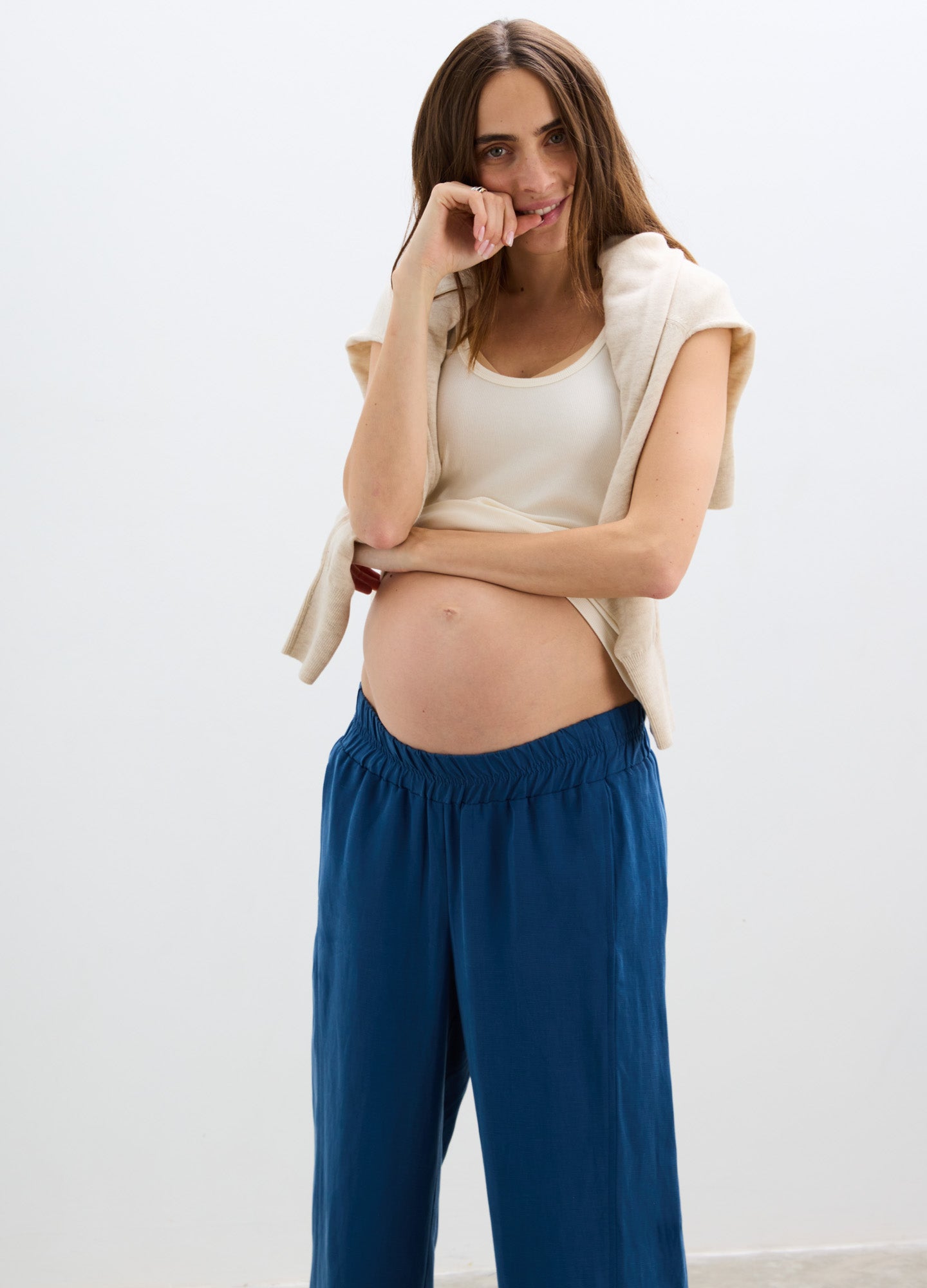 Blue loose-fit maternity pants with elastic waistband worn by pregnant woman paired with white tank top and draped sweater.